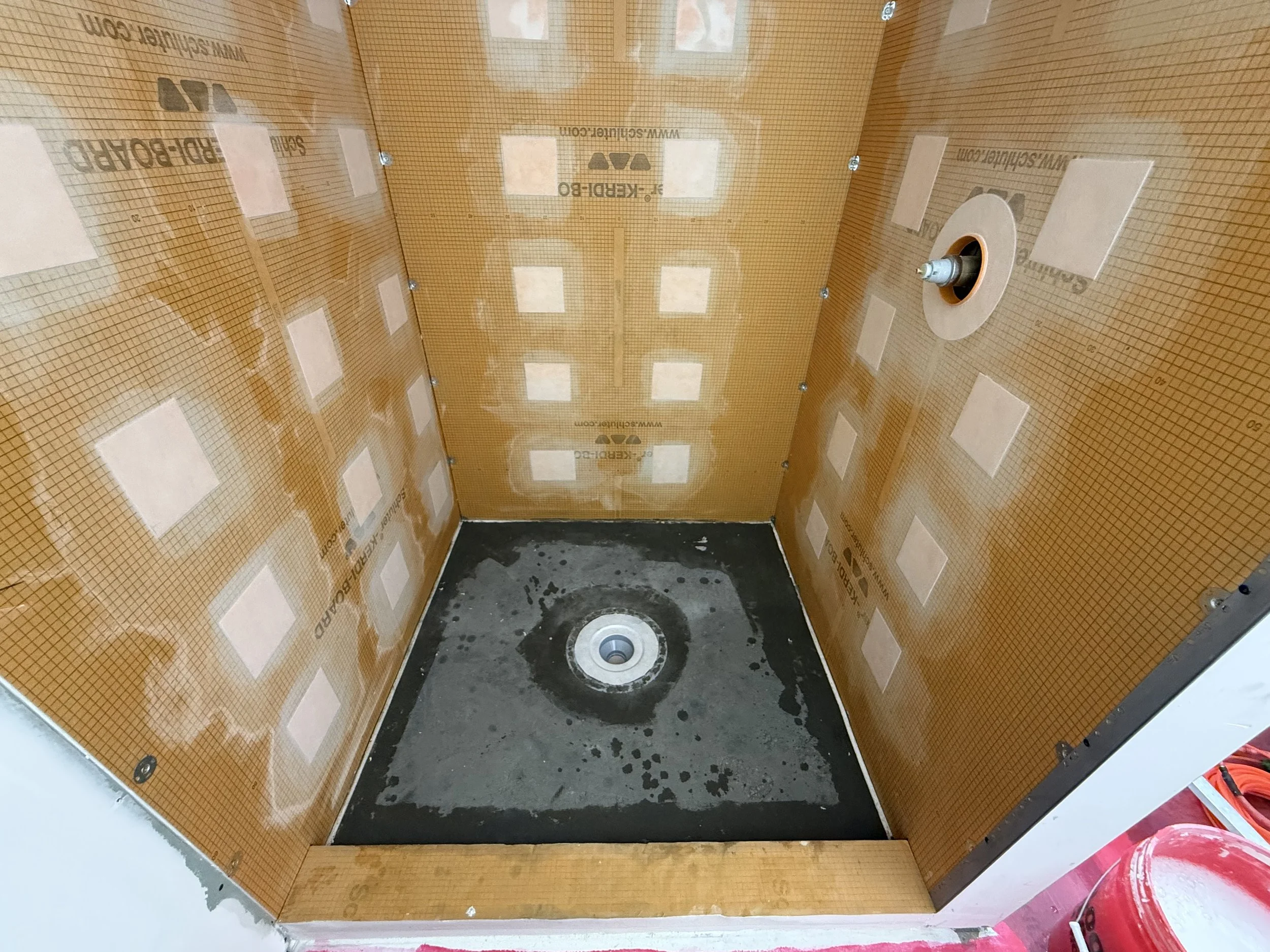 Empty shower stall with orange waterproof wall panels, a drain at the bottom, and plumbing fixtures installed on the wall.