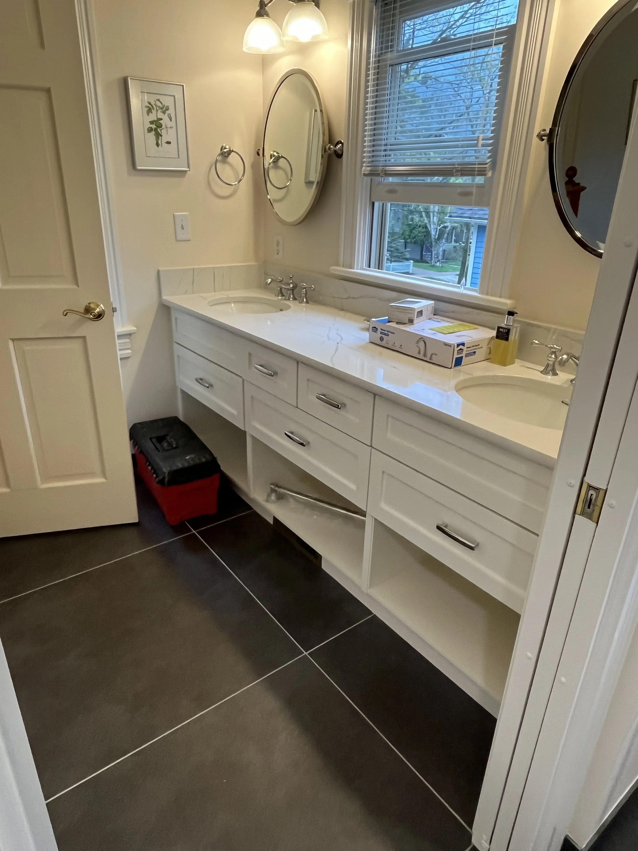 A bathroom with a double sink vanity, large mirrors, and a window with blinds showing a yard outside. The vanity has a marble countertop and cabinetry with drawers. There are two light fixtures above the sinks, and a red toolbox on the dark-tiled flo
