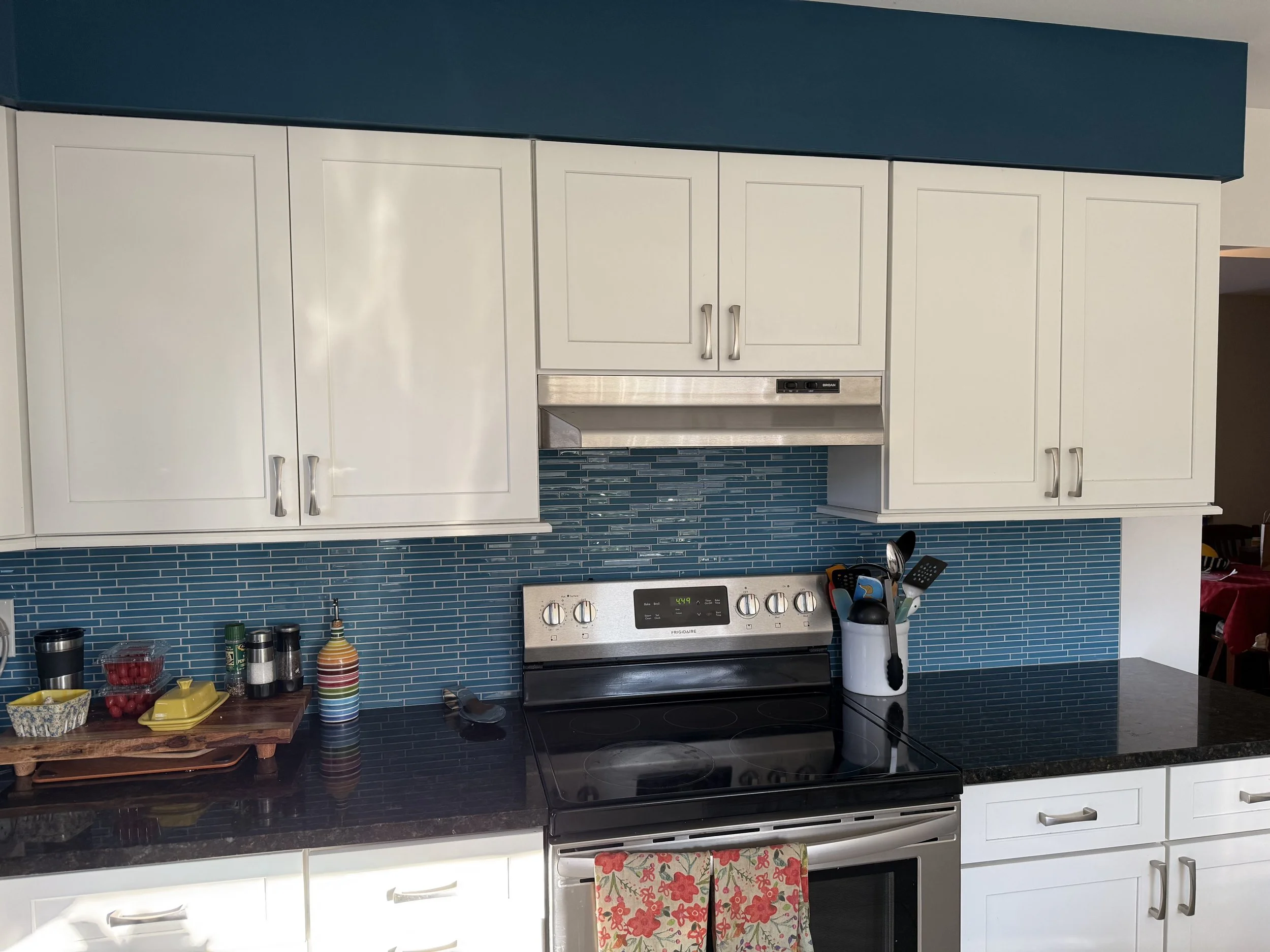 Modern kitchen with white cabinets, black countertop, blue mosaic tile backsplash, stainless steel stove and microwave, and various kitchen utensils and containers.