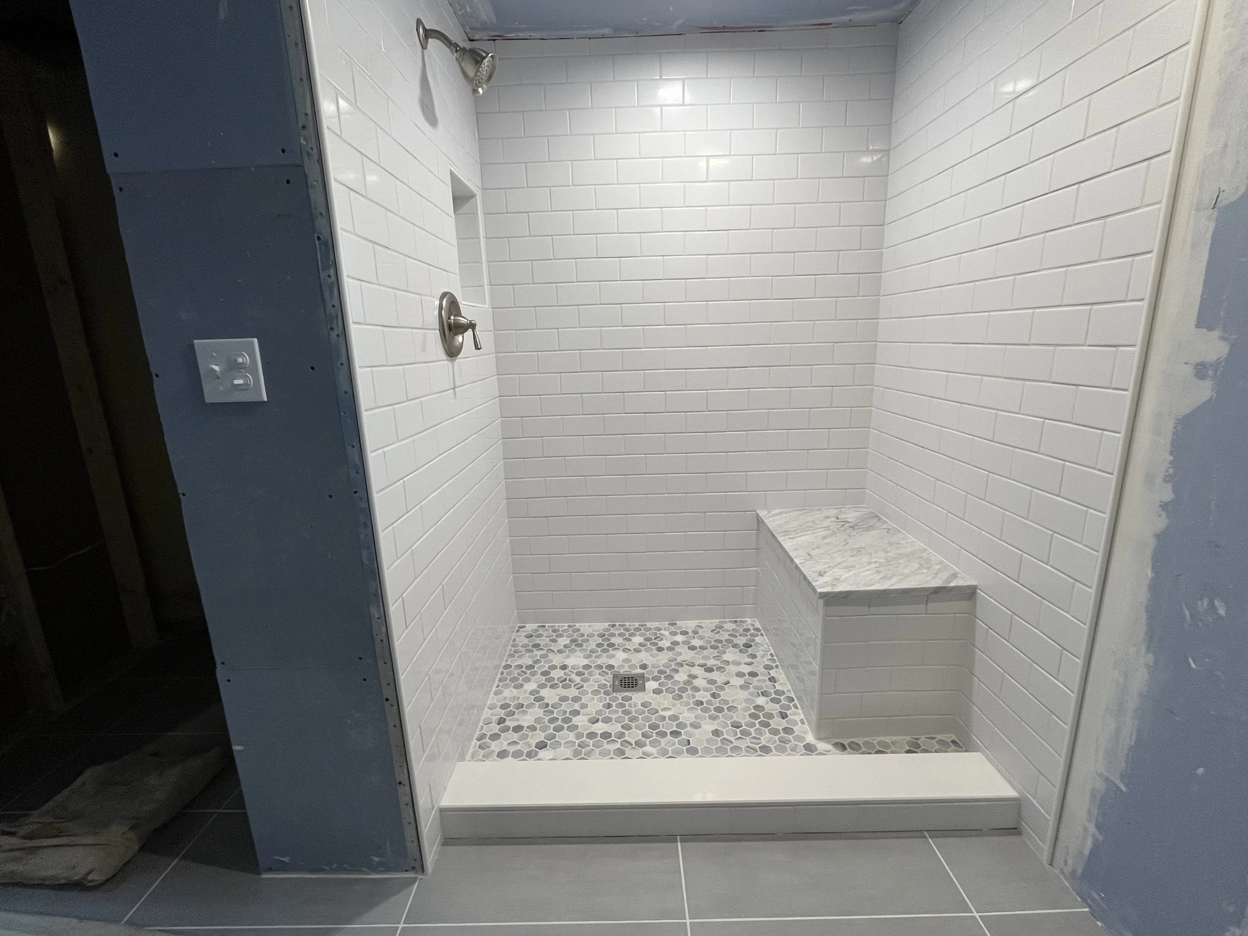 Empty shower stall with white subway tile walls, a marble bench, a small hexagonal tile floor, and a drop-in drain. There is a showerhead and a single handle water control on the wall.