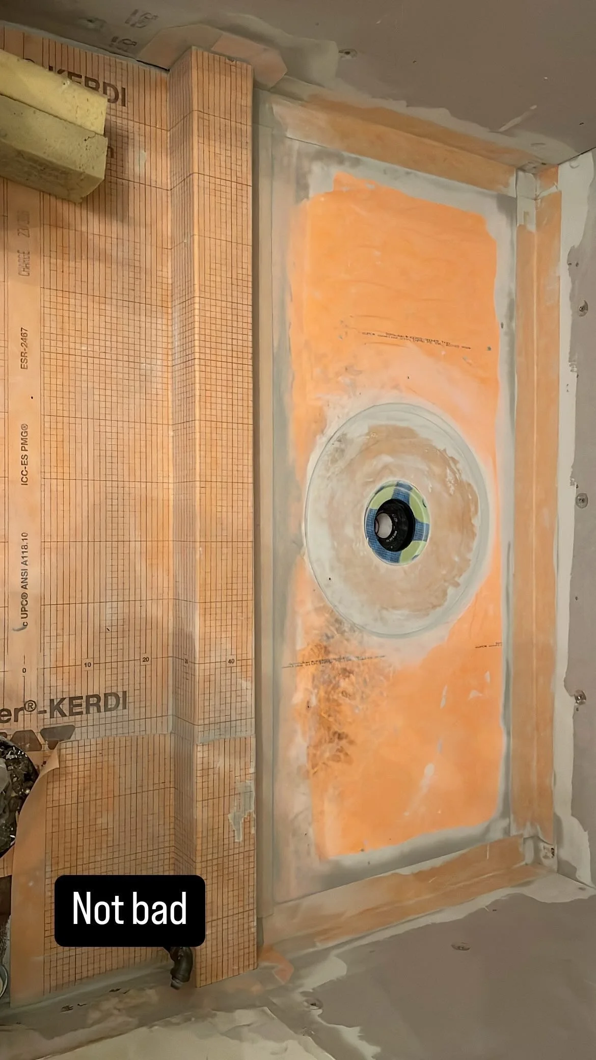 A partially renovated bathroom or kitchen wall with drywall and orange waterproofing compound, a circular hole with a drain, and construction materials like foam and tile spacers. The wall has some unfinished areas and tape around the edges.