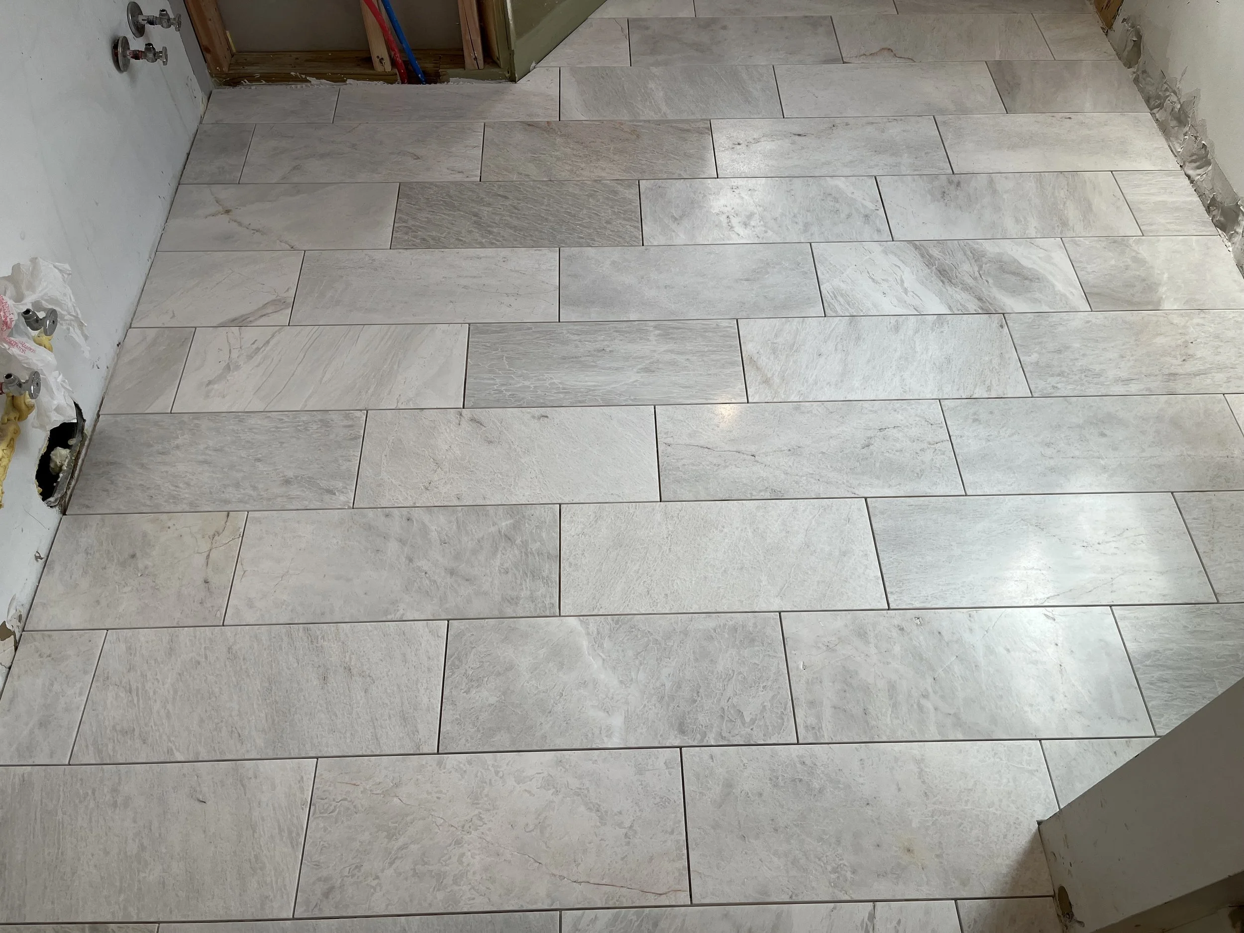 A partially finished bathroom floor with rectangular light gray marble tiles installed, and some construction materials and wall outlets visible in the background.