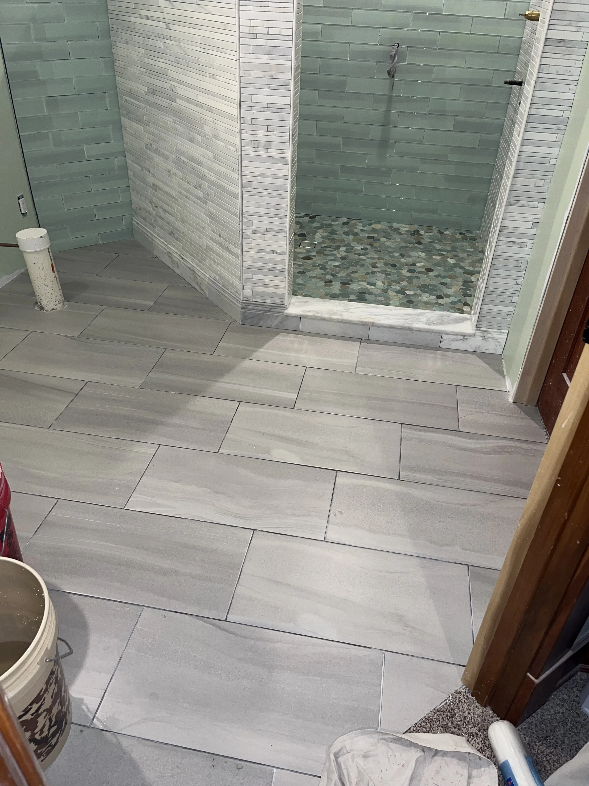 Bathroom with large rectangular light gray floor tiles, a partially visible shower with pebble stone floor and patterned light green and gray tiles on the walls, construction tools and materials nearby.