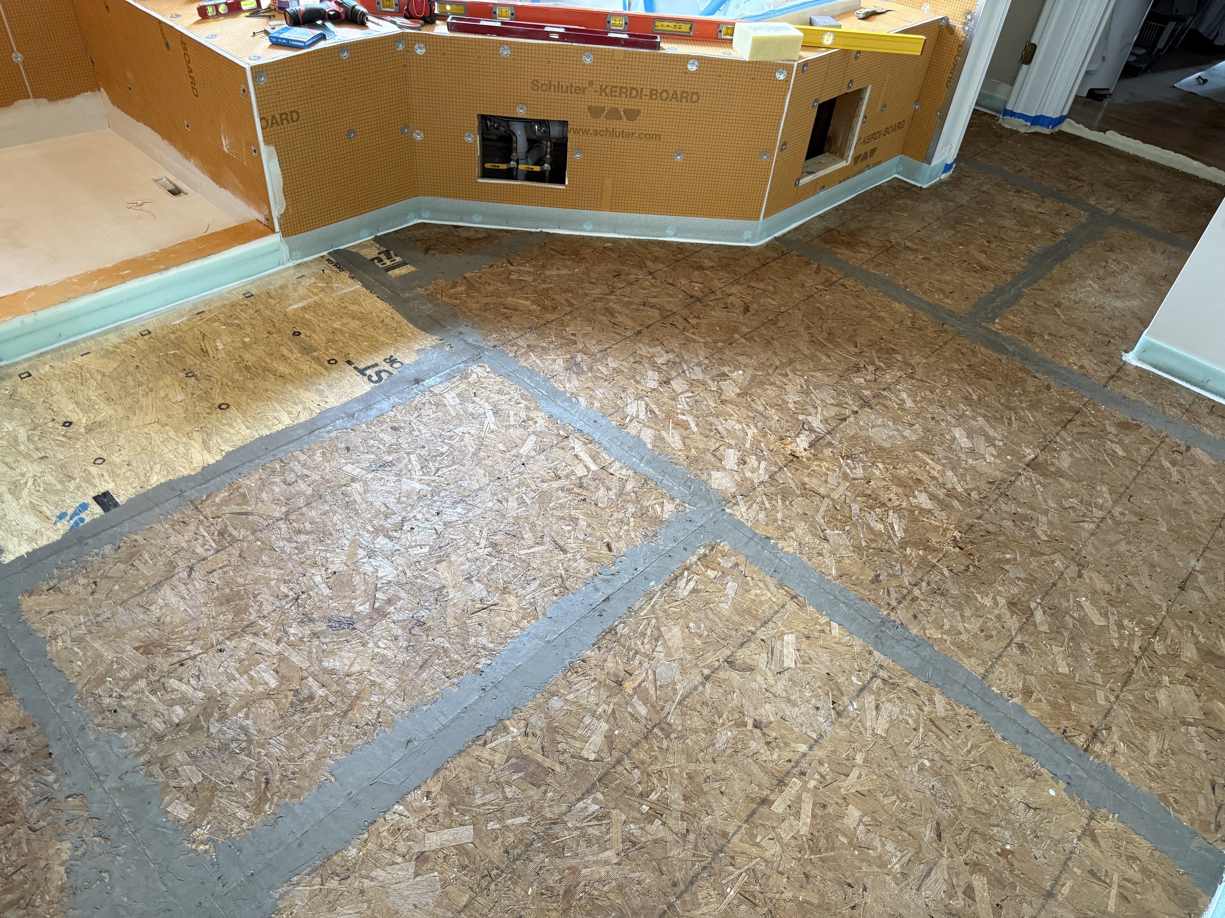 Subflooring installation in progress in a kitchen area, showing oriented strand board (OSB) panels laid out and partially taped with gray sealant; an orange drywall or backer board panel is visible in the background with tools on top.