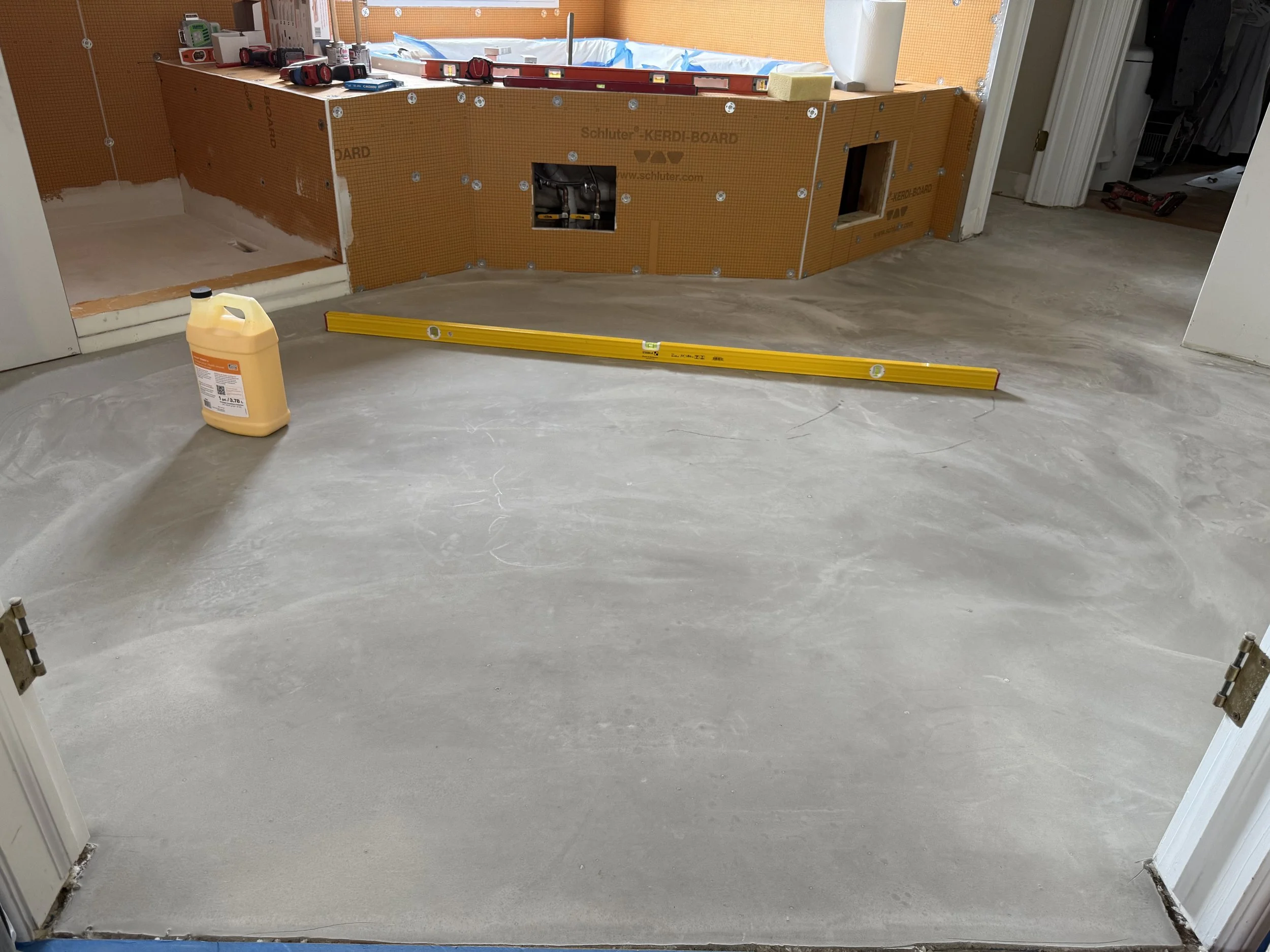 Bathroom under renovation with unfinished floor, a yellow level on the ground, and construction materials including a liquid container and tools on the countertop.