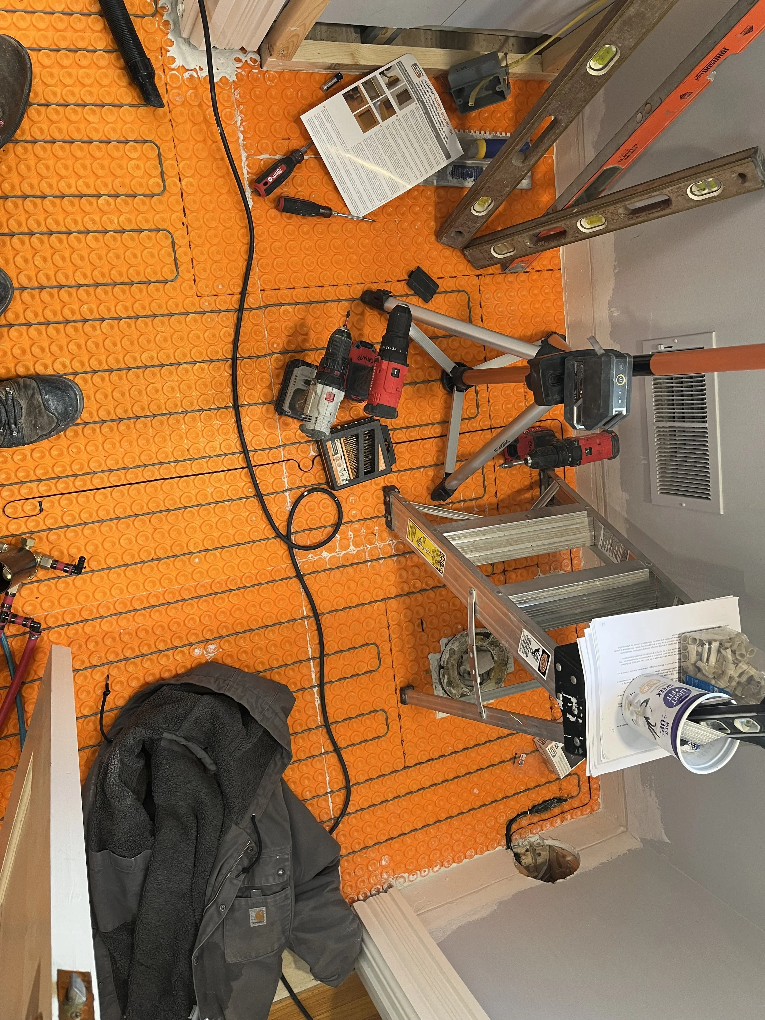 Construction site with orange anti-fatigue mats, power tools, step ladder, and construction materials on a partially finished floor.