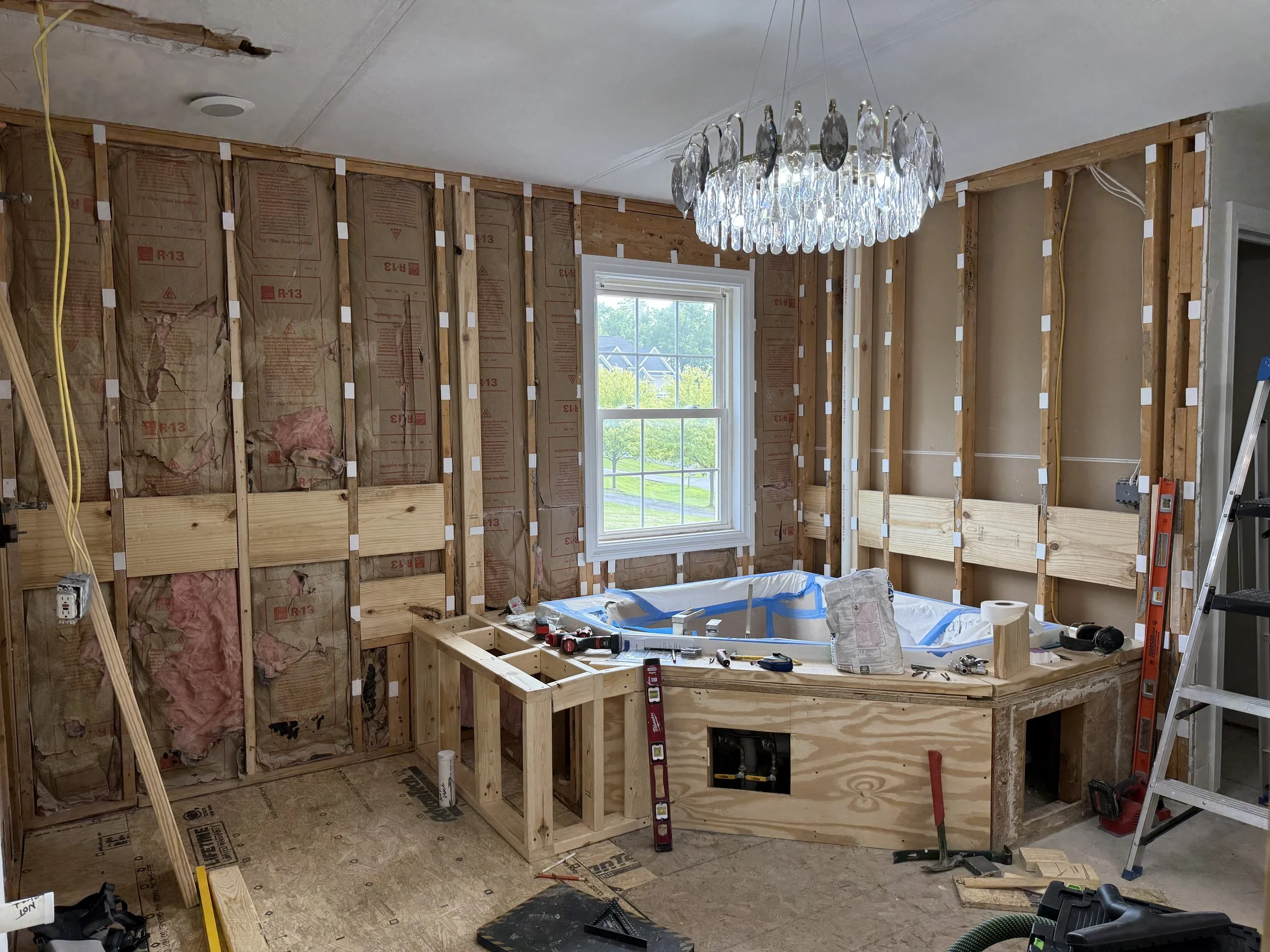 Interior of a room under renovation with exposed wall studs, insulation, a window, a chandelier, and construction tools.