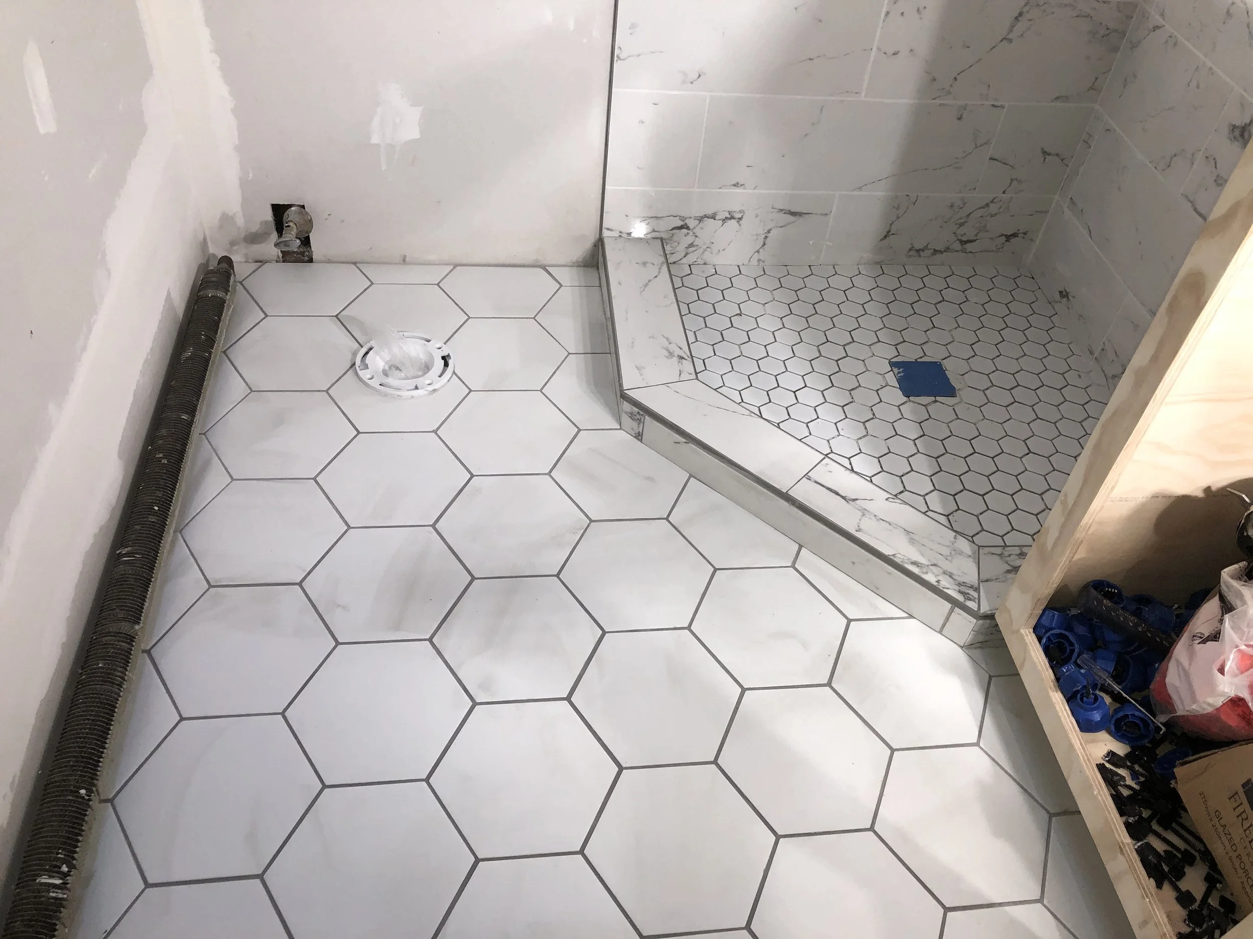 Bathroom with hexagonal white floor tiles, a marble shower area with small hexagonal tiles, and a vent pipe along the wall, under construction.