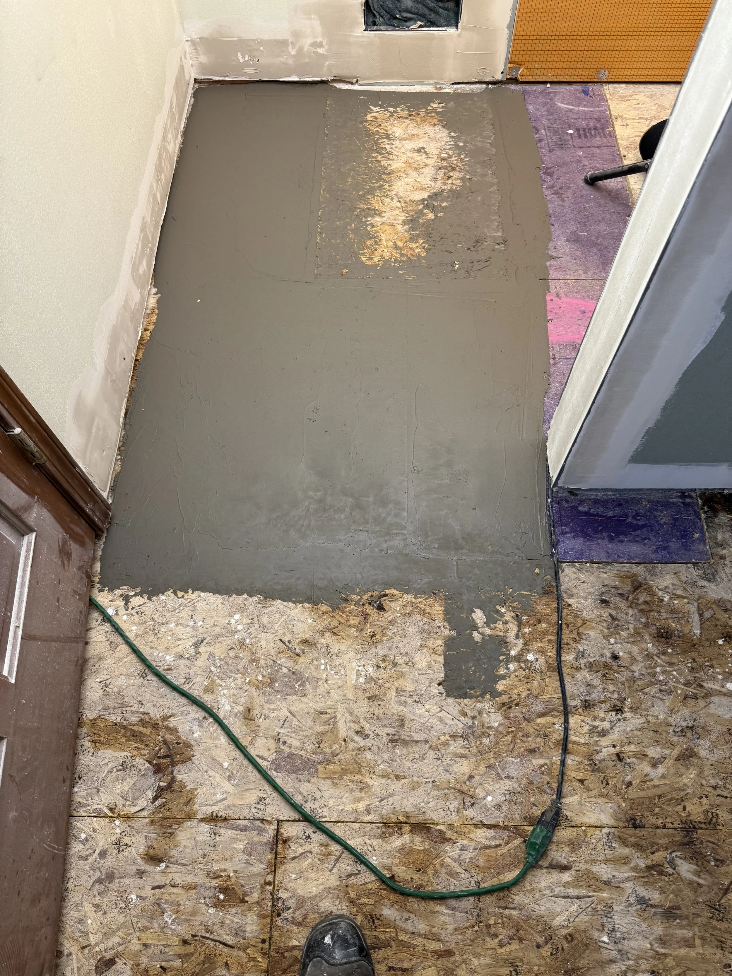 A room under renovation with a concrete floor, some plywood subfloor, and a section of freshly poured gray self-leveling compound. A black electrical cord runs across the floor, and a sock is visible in the bottom center of the image. The walls are p