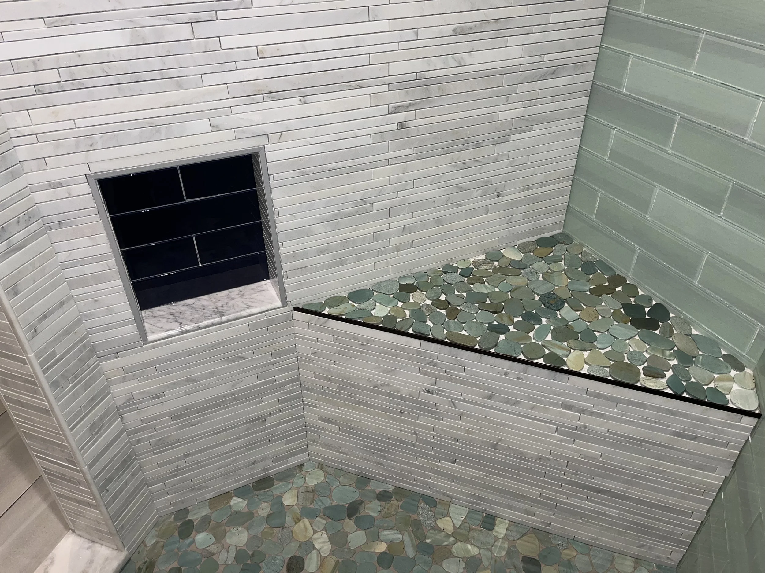 Corner of an indoor space with white tiled walls, a small square window with a marble sill, a separate green tiled wall, and a ledge filled with round multicolored stones.