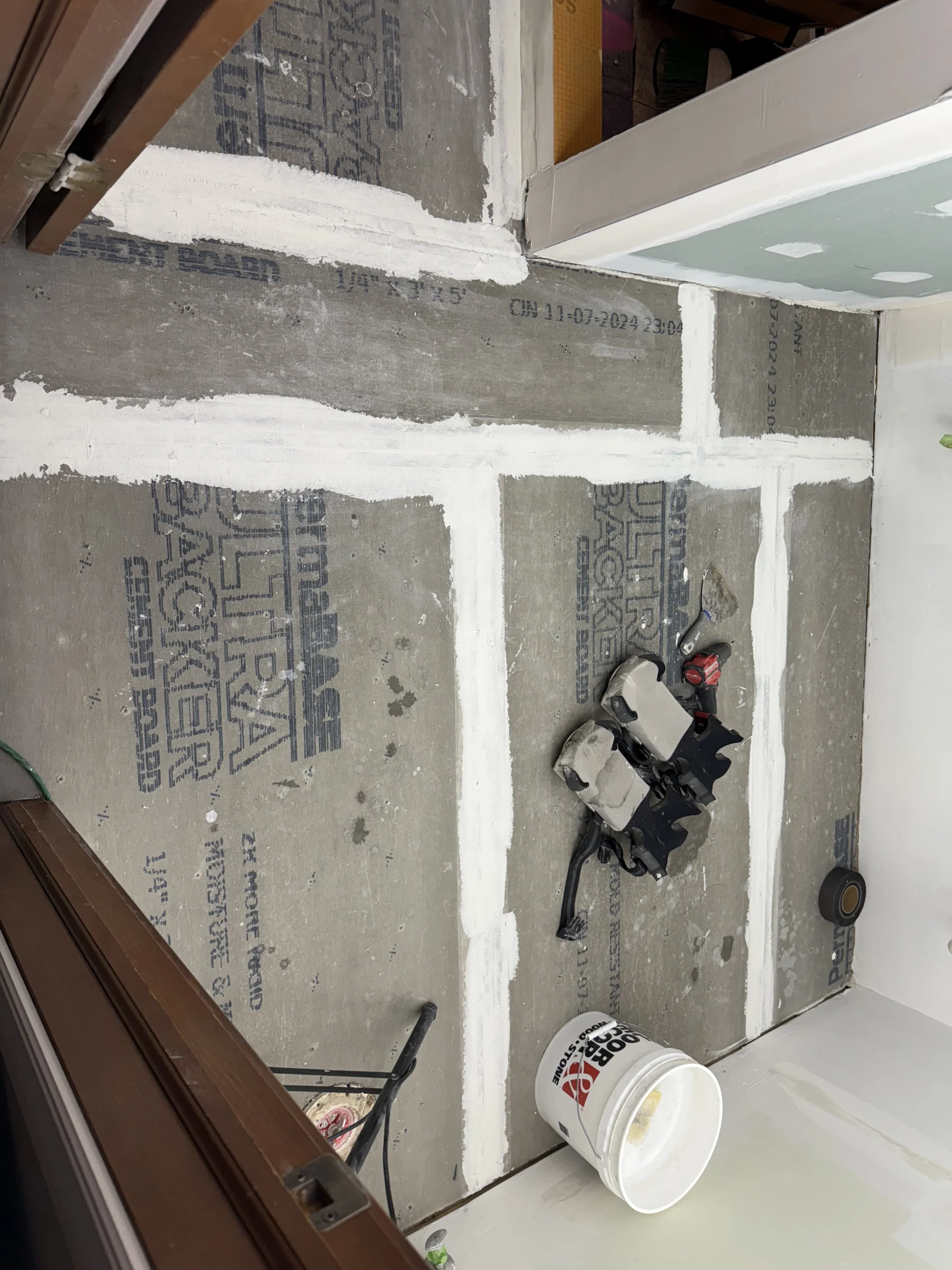 Part of a room under construction with exposed subfloor, white joint compound applied to seams, and construction tools and materials such as a bucket, a power tool, and tape on the floor.