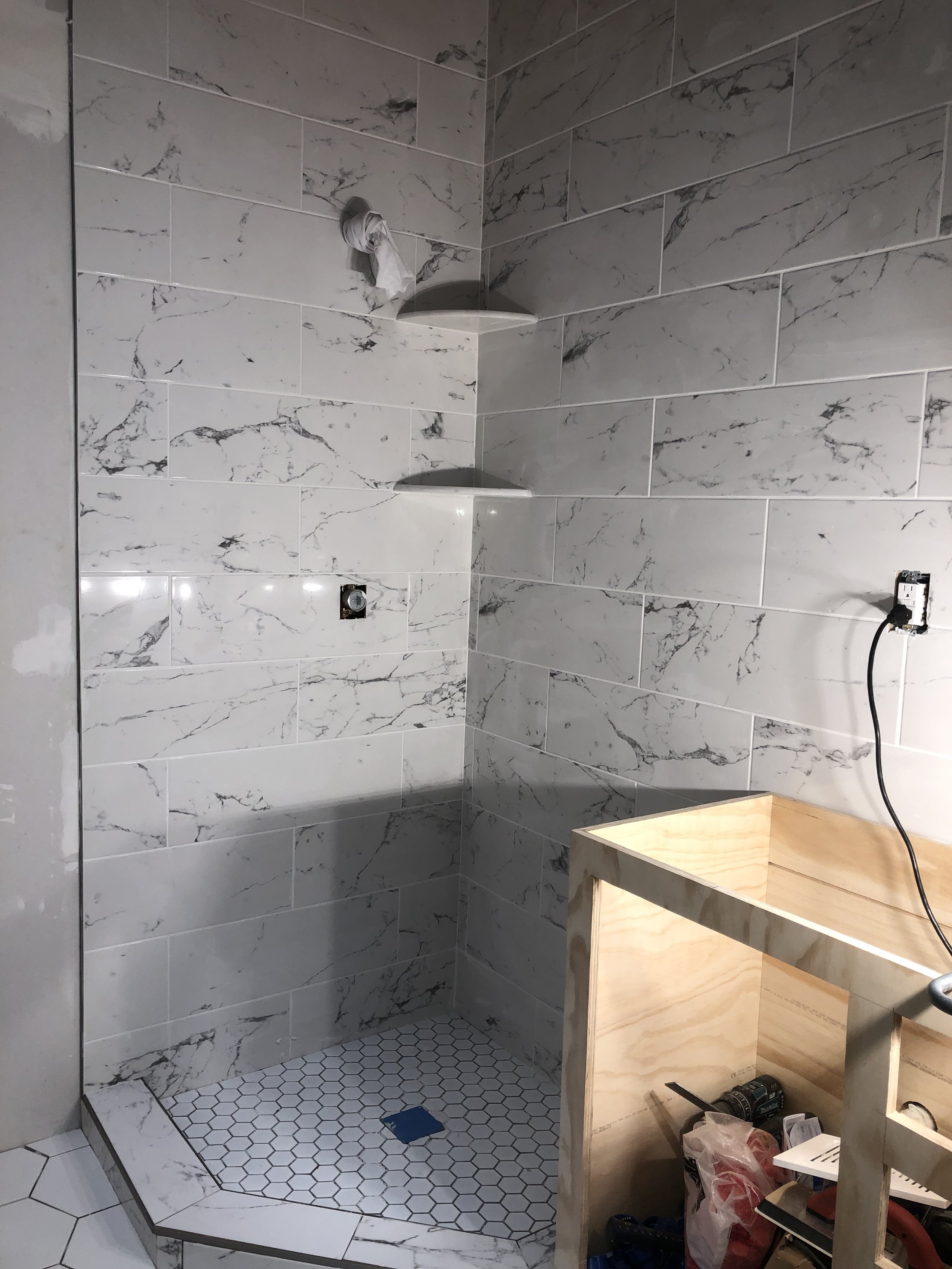 A partially constructed shower area with marble tile walls, a small tiled shower floor, and a wooden cabinet base. There are electrical outlets and wiring visible.