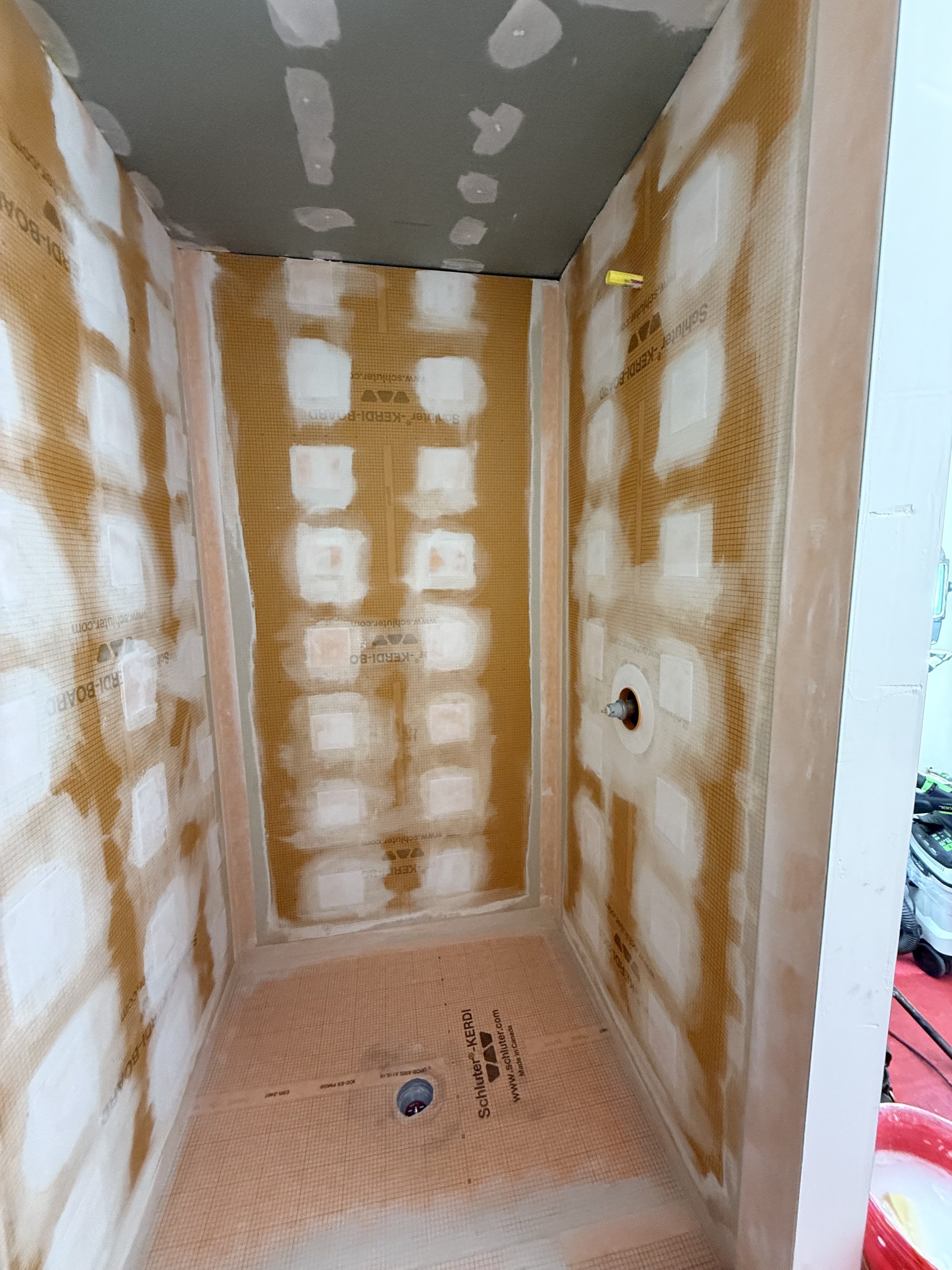 Bathroom under construction with walls and ceiling with drywall panels, some areas taped and mudded, and plumbing pipes visible.