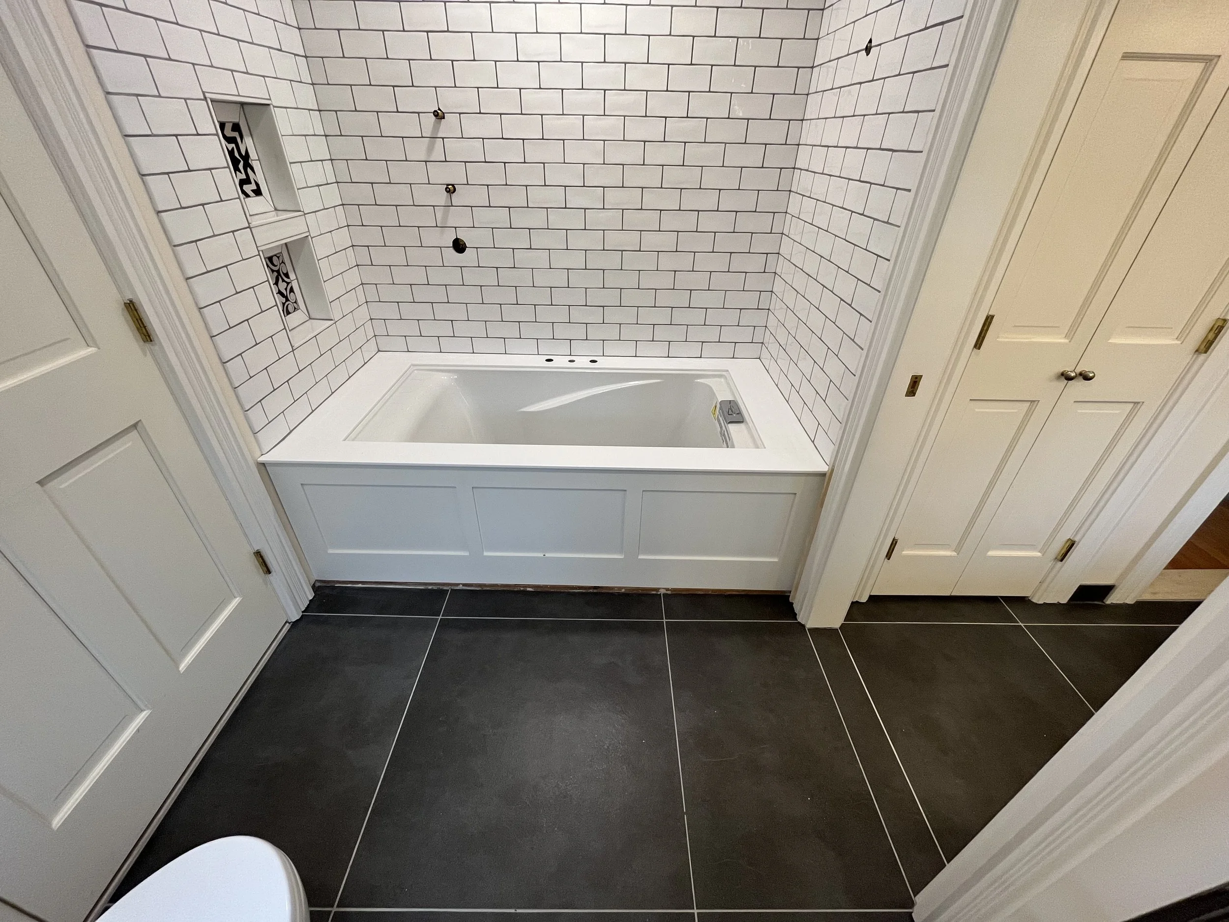 Bathroom with a white bathtub set into a tiled wall with built-in shelves, white cabinets, and black floor tiles.