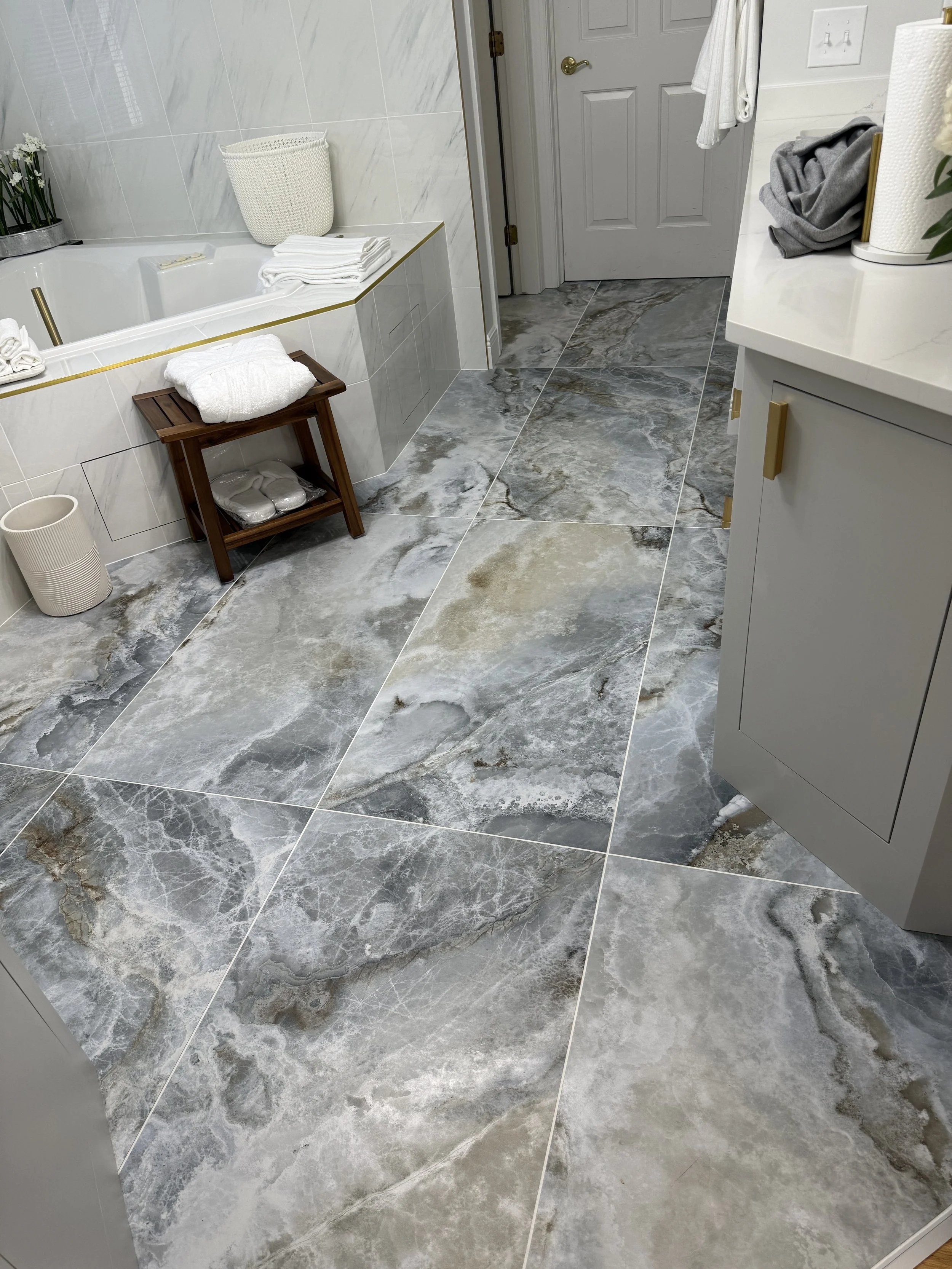 A bathroom with large, marble-patterned gray and beige floor tiles, a corner bathtub with white towels and a basket nearby, a small wooden stool with a towel on top, a beige vase, and a gray vanity with gold handles.