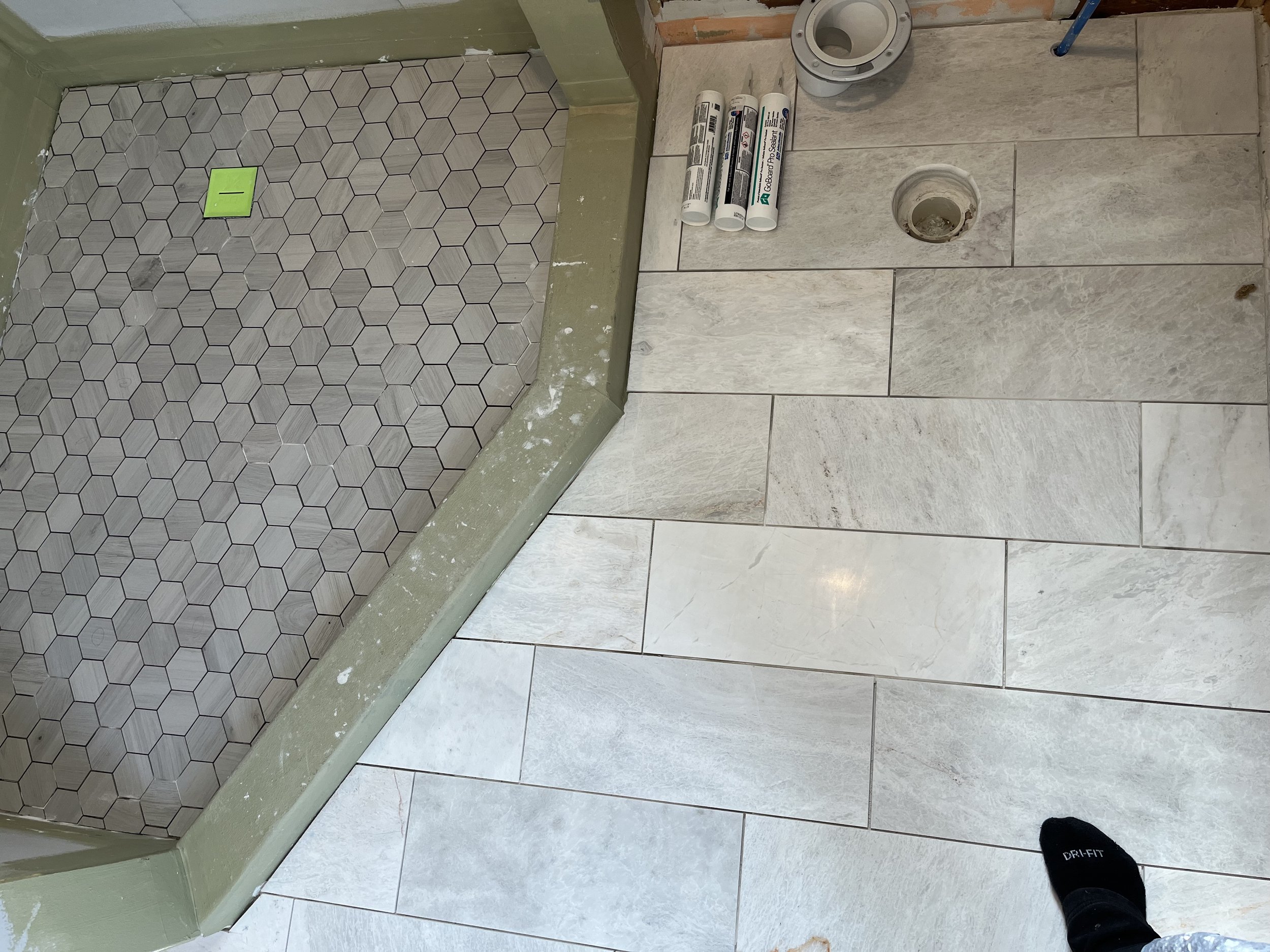 Bathroom floor with large light-colored tiles and a shower area with hexagonal tiles. A green painter's tape is on the shower floor. Toilets and plumbing fixtures are visible, along with four cans of construction sealant and a pipe opening on the flo