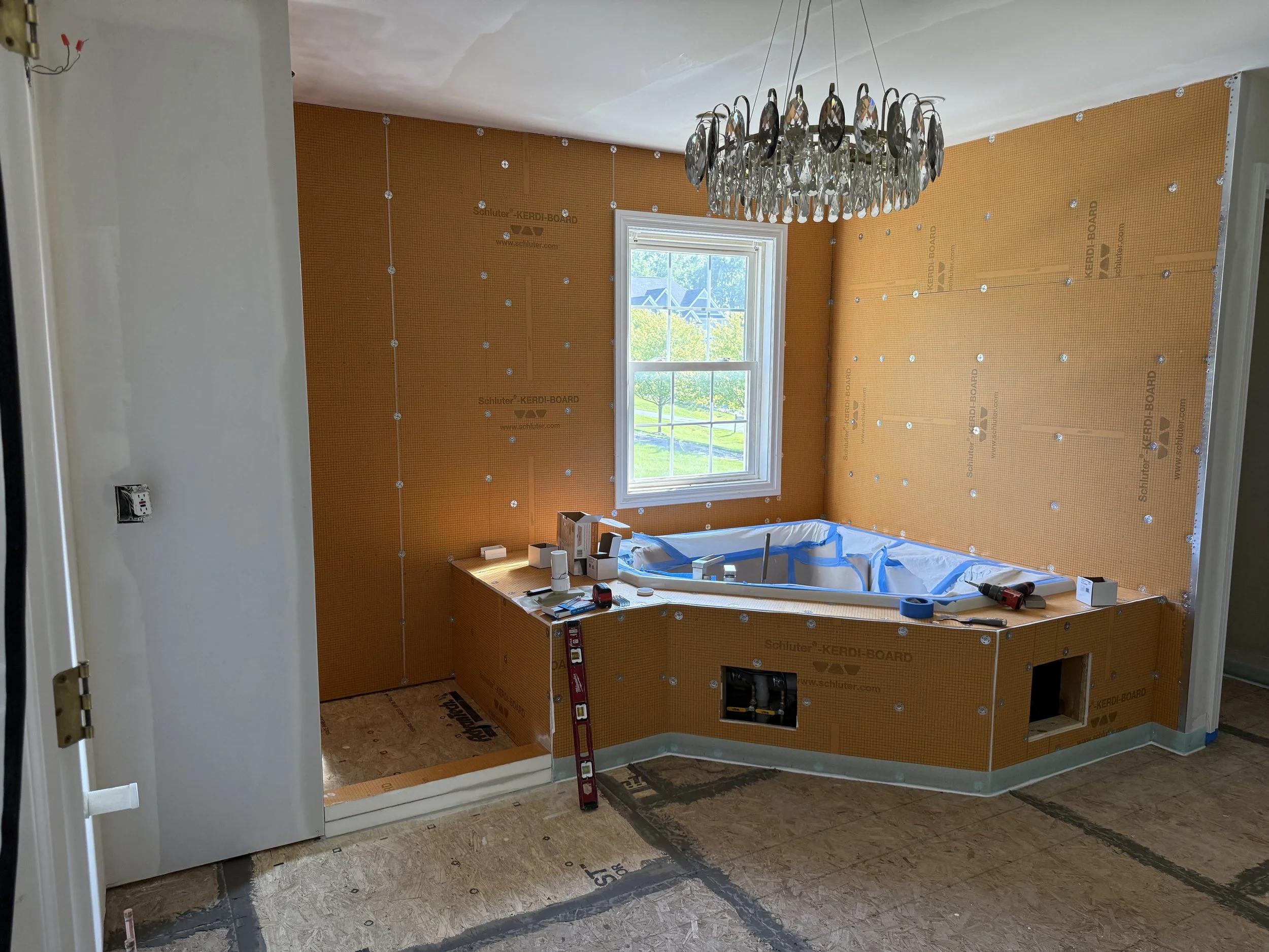 A partially renovated bathroom under construction with a window, orange drywall, and a built-in bathtub covered with blue painter's tape. There is a chandelier hanging from the ceiling and construction tools and materials on the tub and floor.