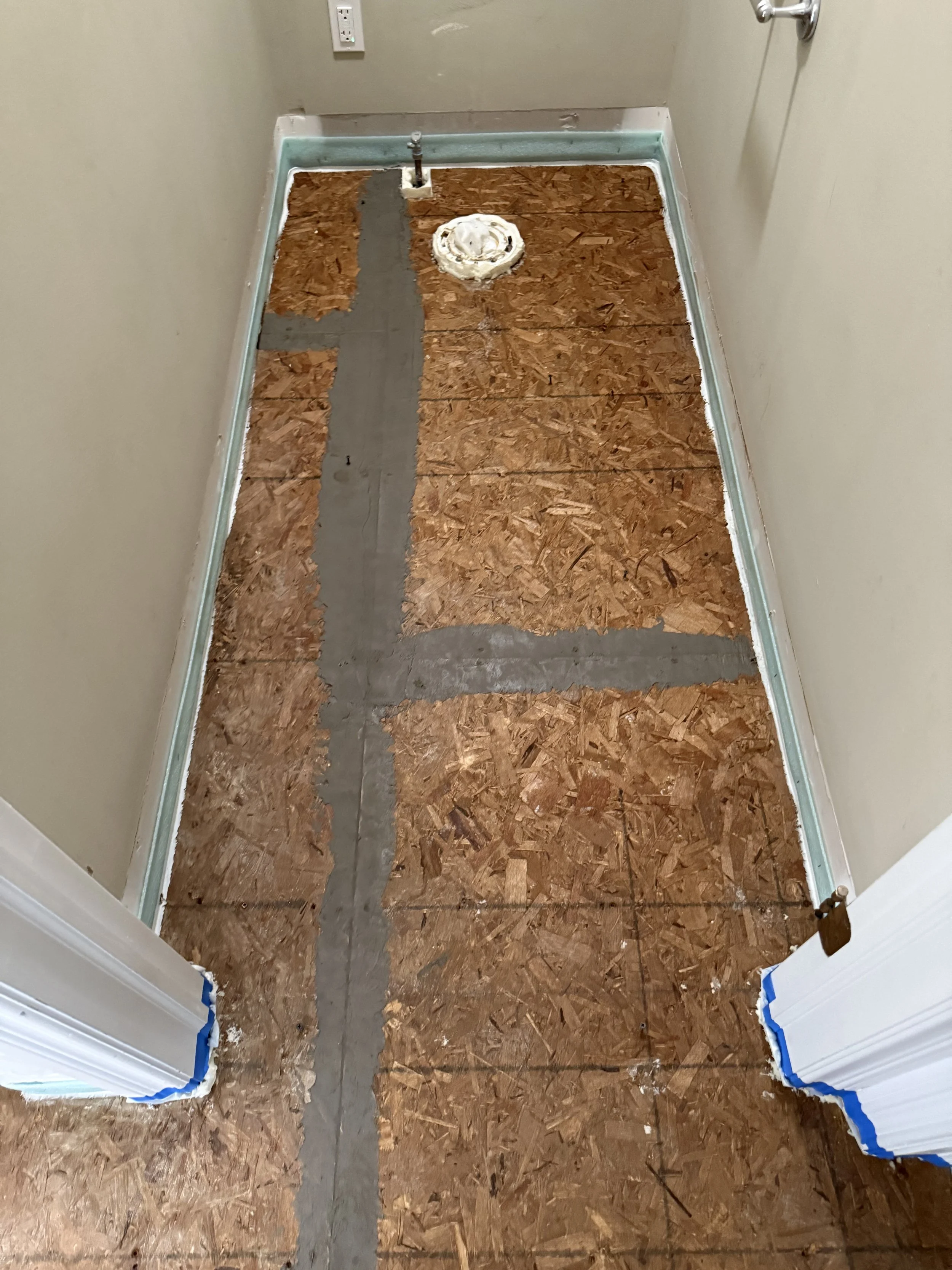 Floor stripped to subfloor with visible sawdust, with plumbing and vent pipe in the process of renovation, surrounded by cream-colored walls and white trim, with doorway ready for flooring installation.