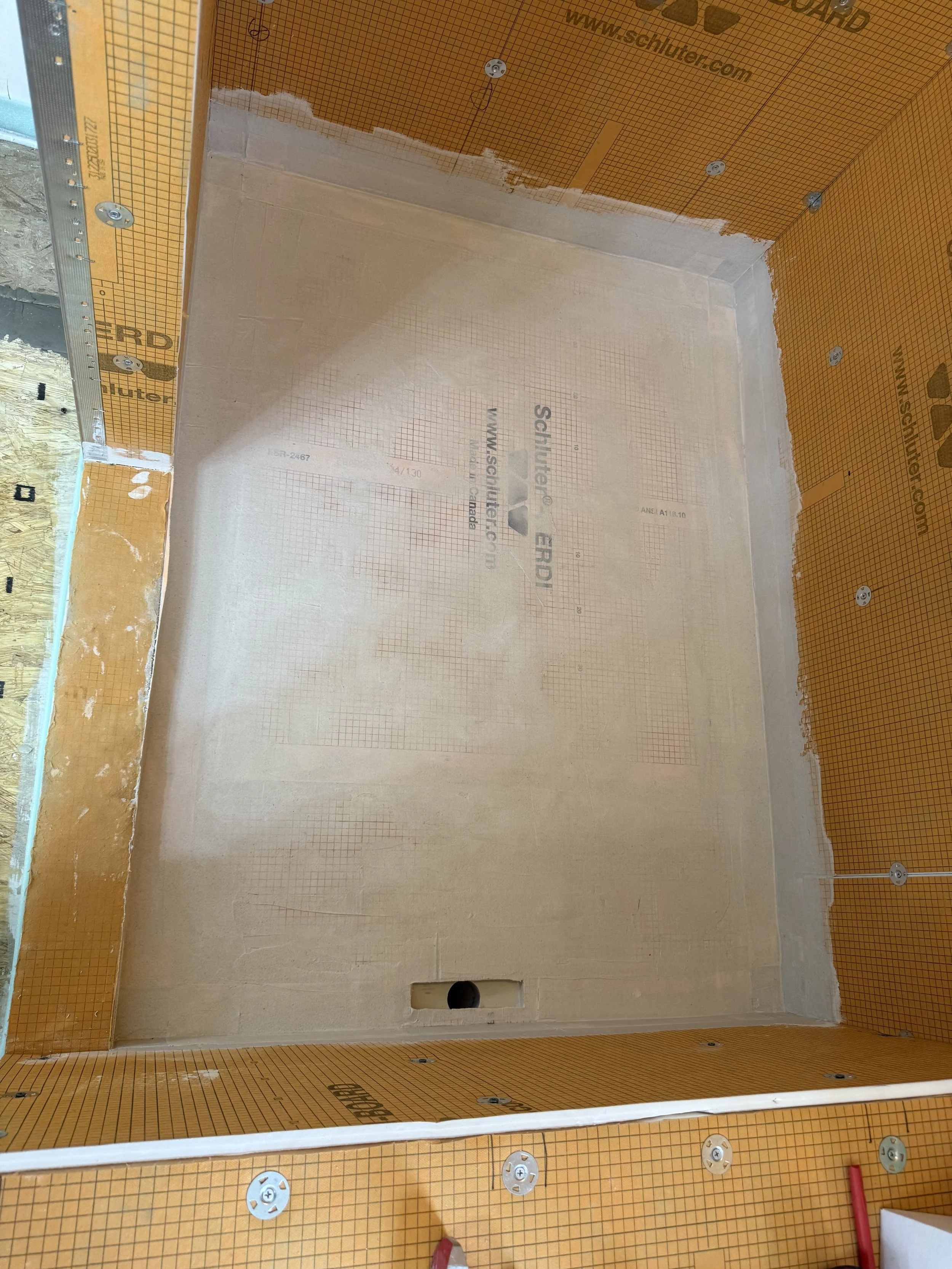 Interior of a bathroom or shower stall under construction, showing yellow waterproofing membrane on walls and floor, with some visible plumbing pipe and electrical wiring.