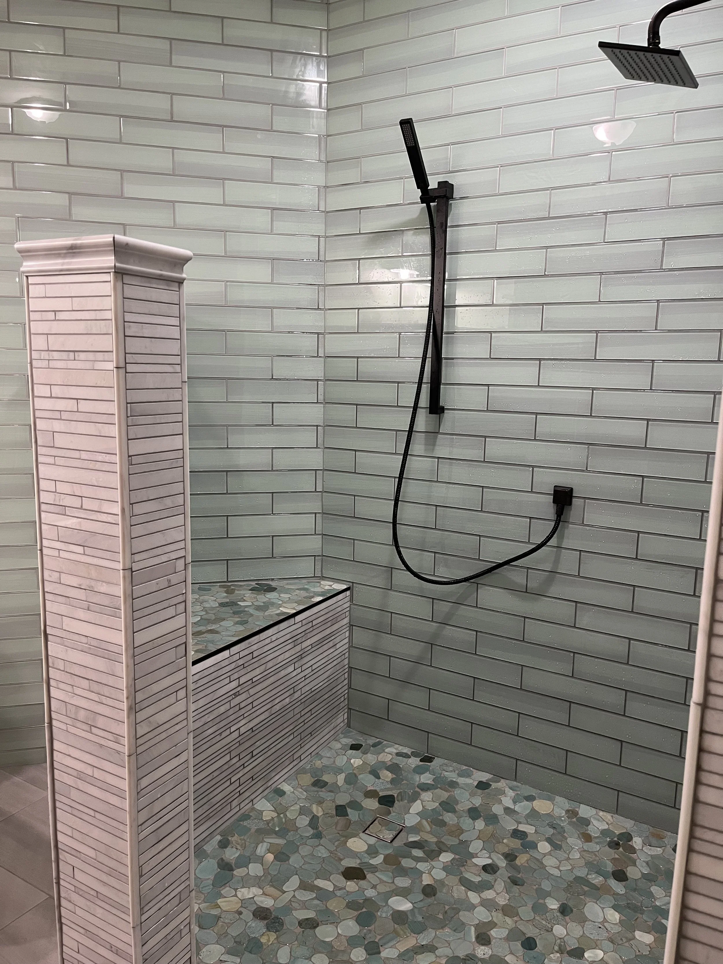 A modern walk-in shower with green subway tiles on the walls, pebble tiles on the floor, a mounted square rainfall showerhead, a black handheld shower wand on a vertical bar, and a vertical white brick accent column.