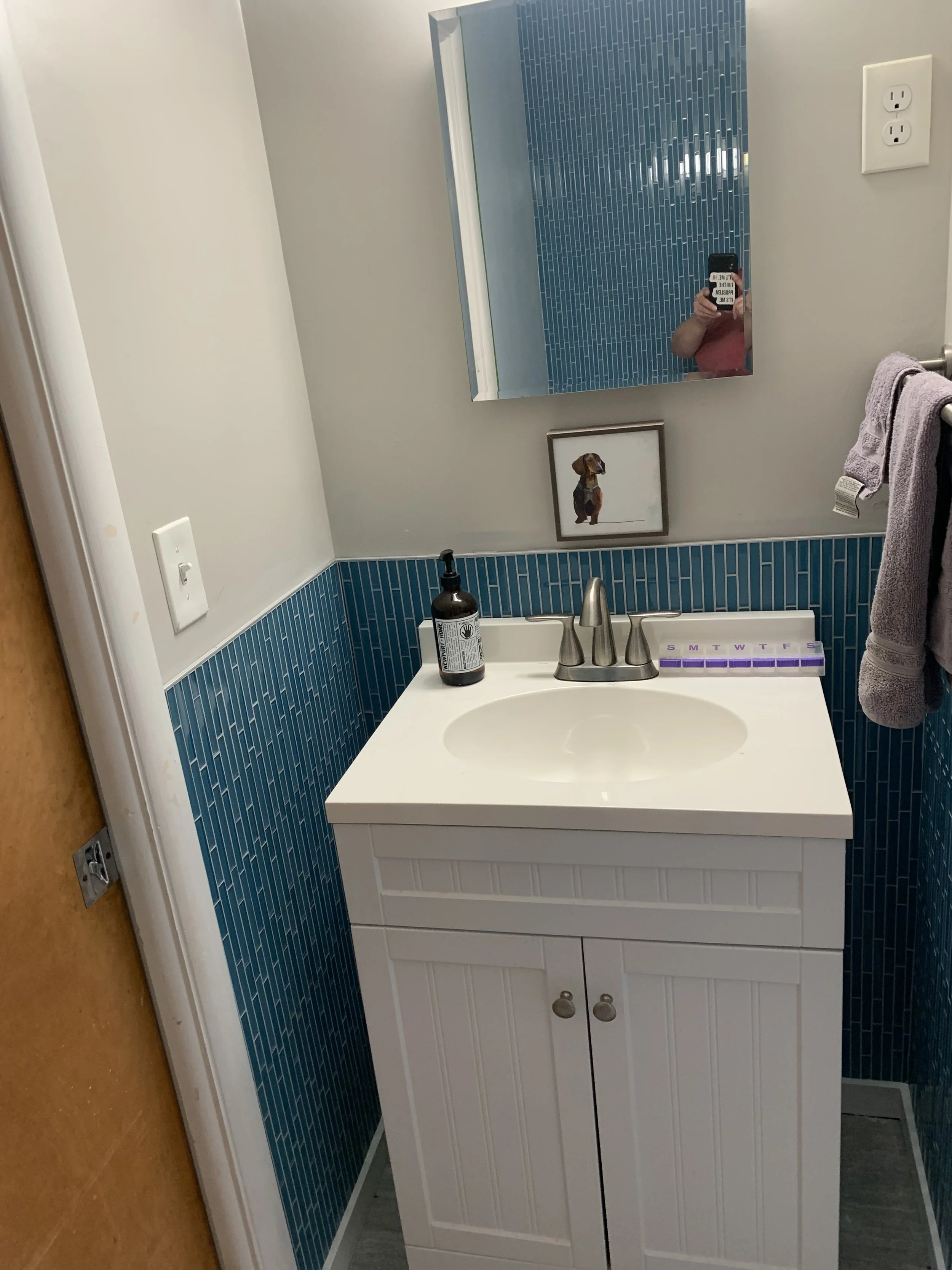A small bathroom vanity with a white cabinet and a oval sink, a soap dispenser, a purple weekly pill organizer, a mirror, a framed picture of a dog, a purple towel on a rack, and a wall outlet.