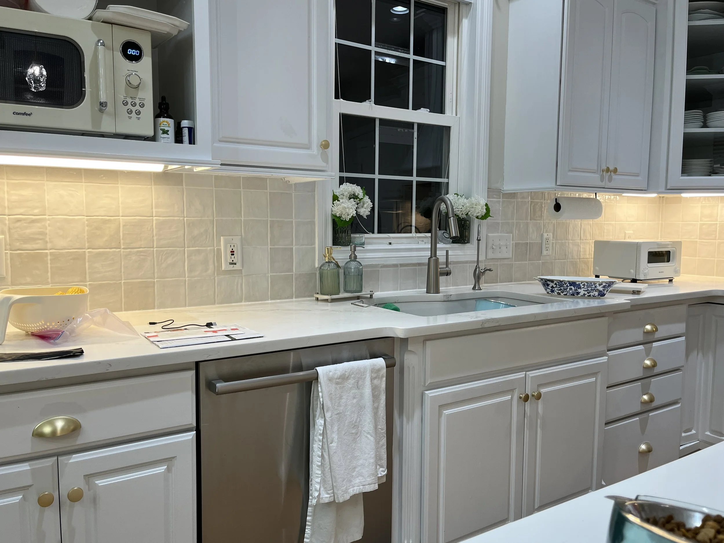Kitchen with white cabinets, a beige tiled backsplash, a white marble countertop, a sink, a window with white panes, and various kitchen appliances and items, including a microwave, a toaster, and flowers in vases.