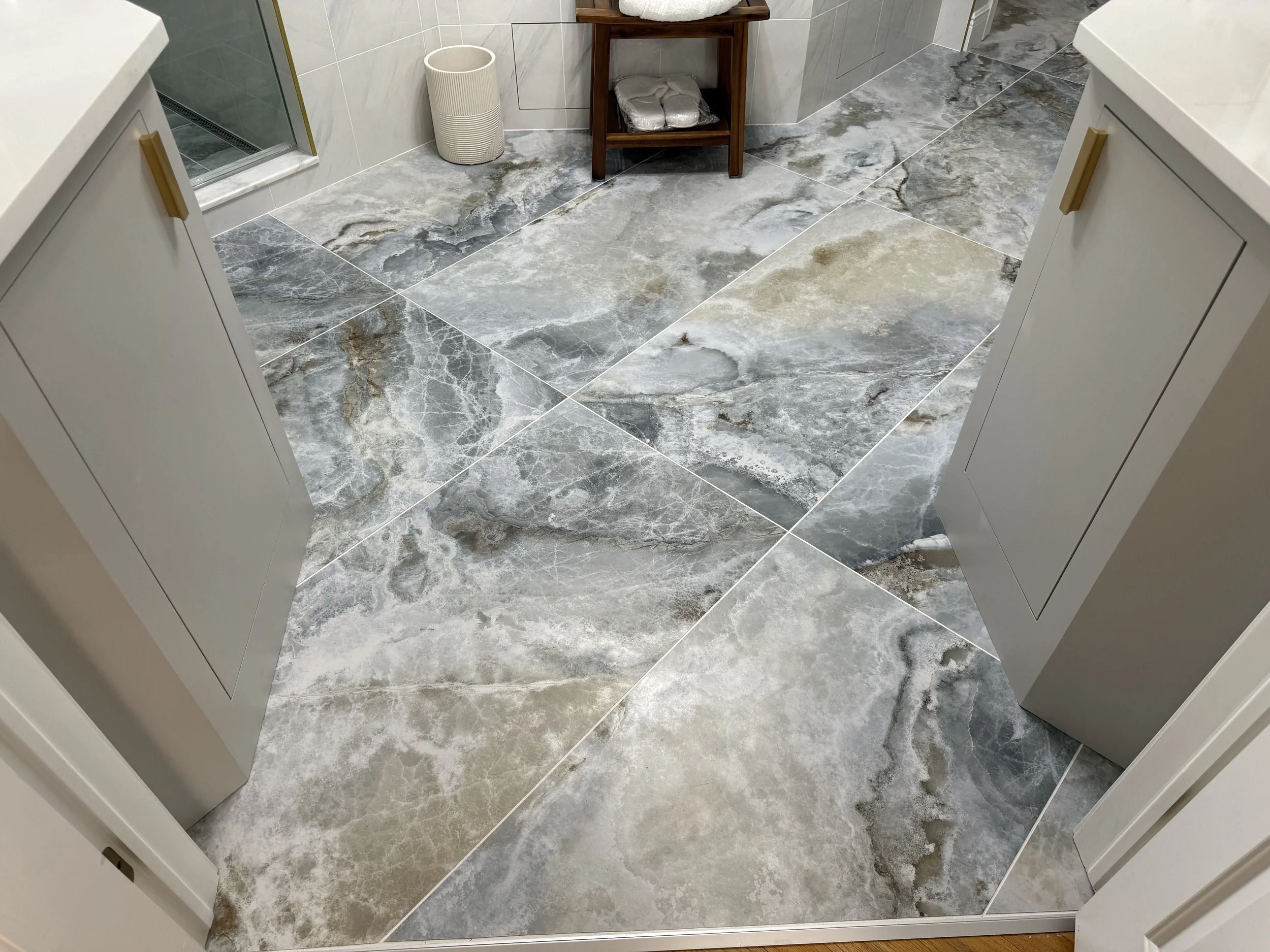View of a bathroom floor with large, marble-like tiles in shades of gray, beige, and white. Two white vanities with gold handles are on either side of the image. In the background, a small wooden shelf holds towels and a white basket, and a white was