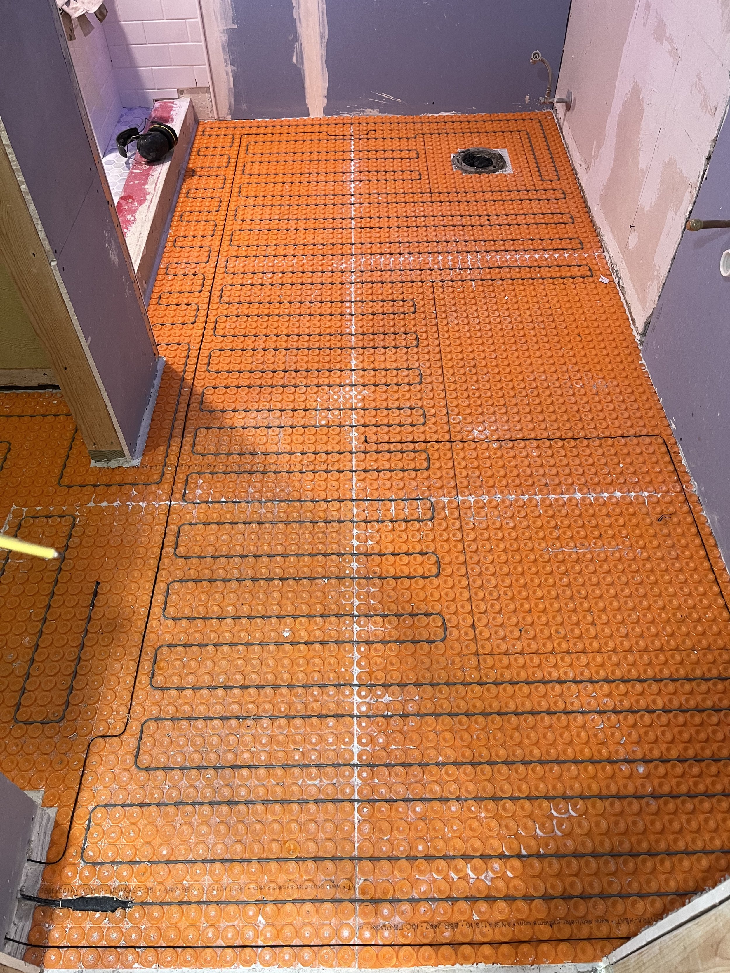 Installation of orange electric radiant floor heating mats in a bathroom under concrete floor, with a drain in the corner and ongoing wall construction.