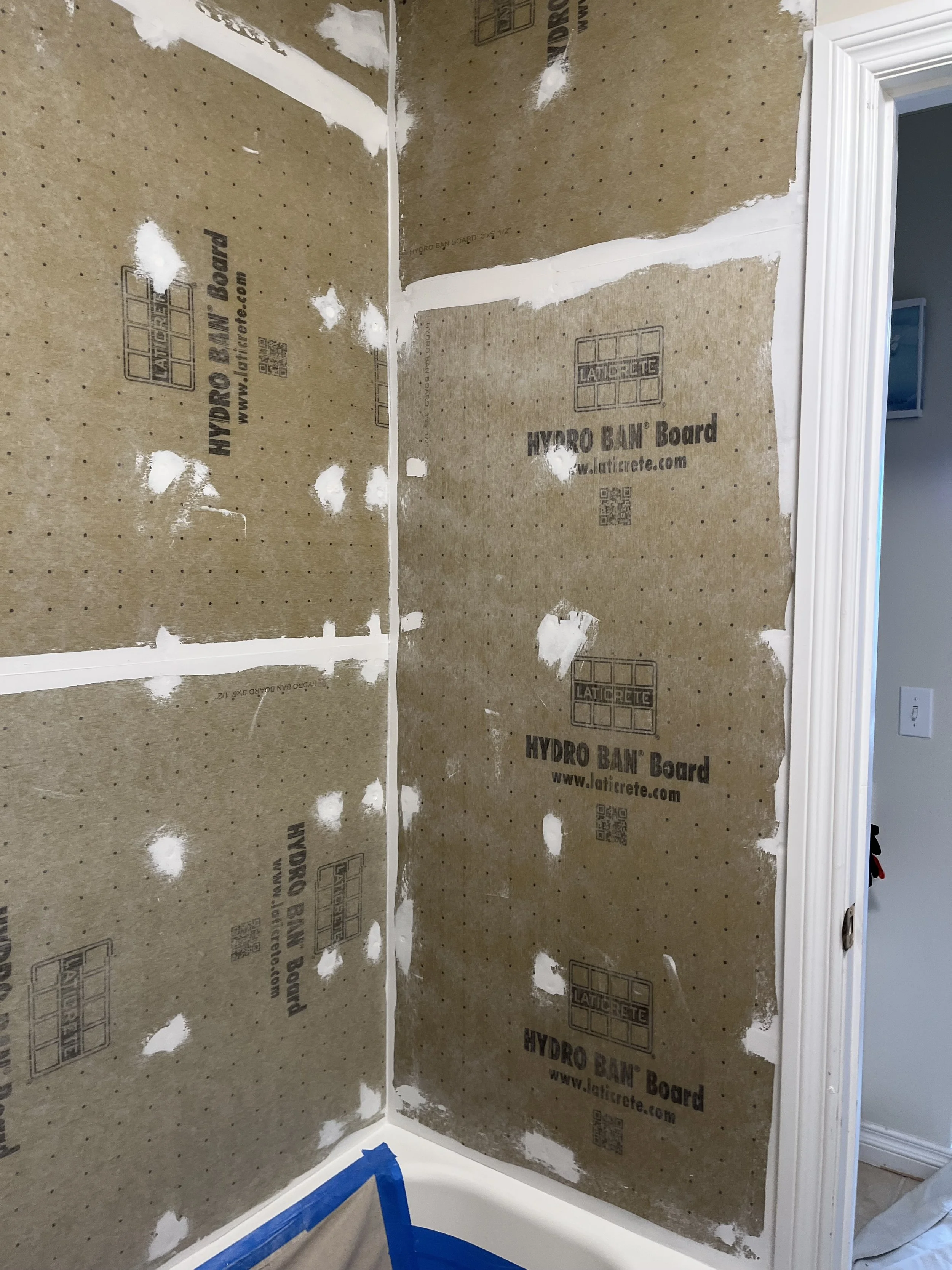Bathroom corner under construction with drywall panels, joint compound, and blue painter's tape on the baseboard.