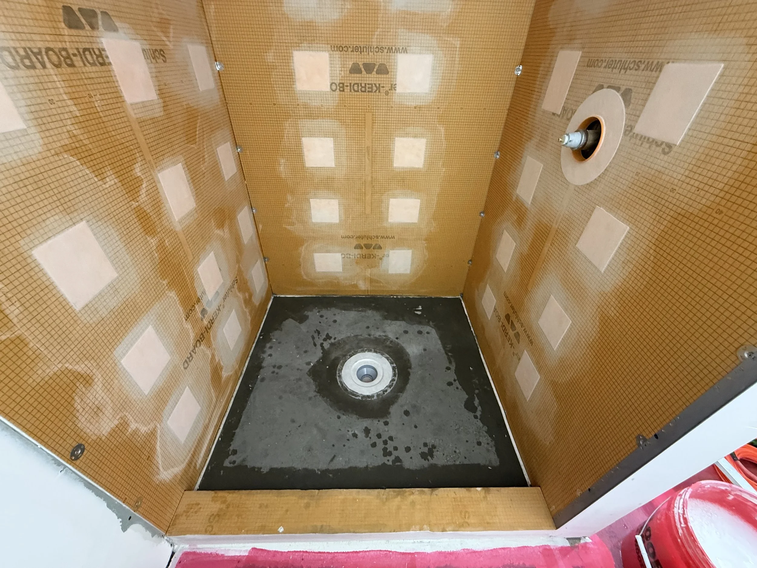 The inside of a small, unfinished bathroom shower area with orange waterproofing membrane on the walls, a drain in the center of the concrete floor, and plumbing fixtures for a shower head on the wall.