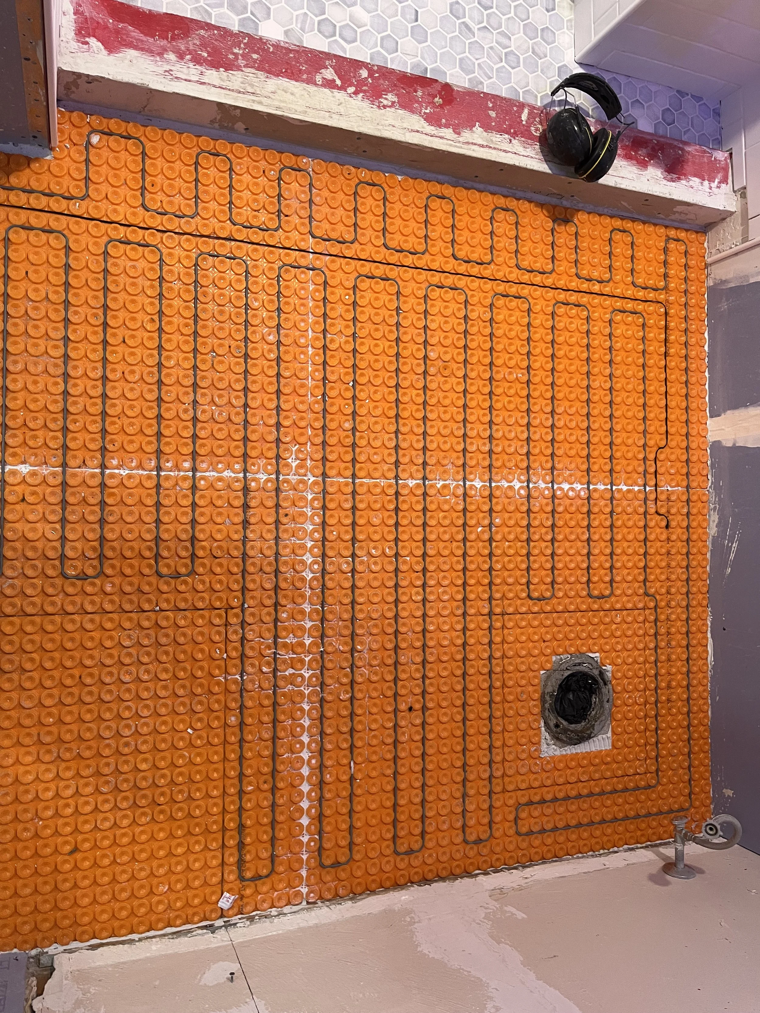 A floor with orange plastic tiles and underfloor heating pipes visible, with a hole for plumbing and a pair of black ear protectors hanging on the wall above.