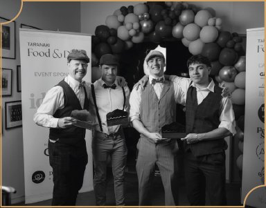 Four men dressed in vintage attire holding food trays at an event with balloon decorations and signage in the background.