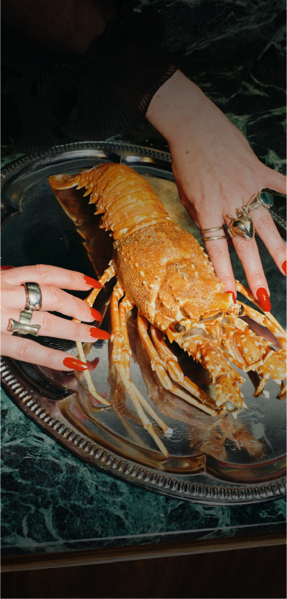 A person with red manicured nails and multiple rings is holding a cooked lobster on a silver platter.