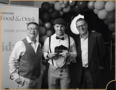 Three men in formal attire standing in front of a balloon arch at a food and drink event, with one holding a black folder or plaque.