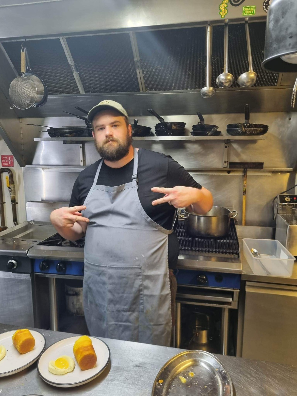 A man with a beard wearing a black shirt, gray apron, and cap standing in a commercial kitchen making peace signs with both hands while posing in front of a stove with pans and kitchen utensils hanging above. Plates with food, including spring rolls and fried eggs, are on the counter in front of him.