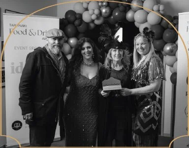 Four people standing together at an event, two women holding an award, with balloons and event signage in the background, in black and white.