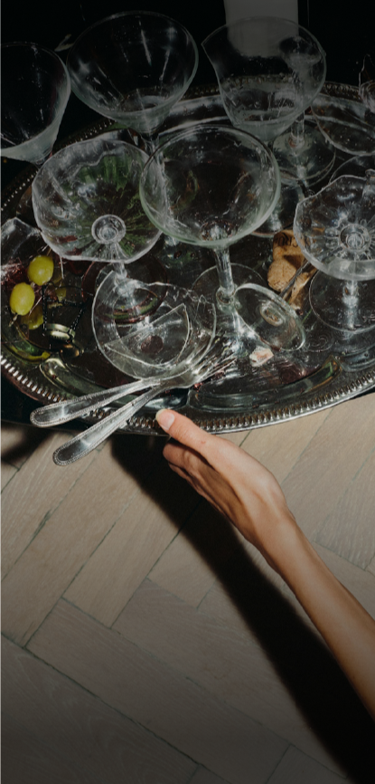 A person holding a large silver tray with multiple empty wine glasses and some grapes and snacks on a wooden floor.