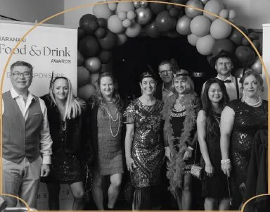 Group of eight people at an awards event, standing in front of a balloon arch and a backdrop with text related to food and drink awards.