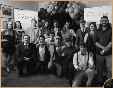 Group of people at a celebration or event, standing and sitting in front of a decorated backdrop with balloons and banners.