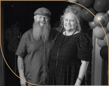A man with a beard and a woman with curly hair smiling, standing together at an event with balloon decorations in the background.