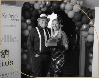 A man and woman are standing together at an event with a balloon arch behind them. The man is wearing a hat, glasses, and a shirt with suspenders. The woman is dressed in a shiny, patterned dress and holding a box. They are smiling for the photo.