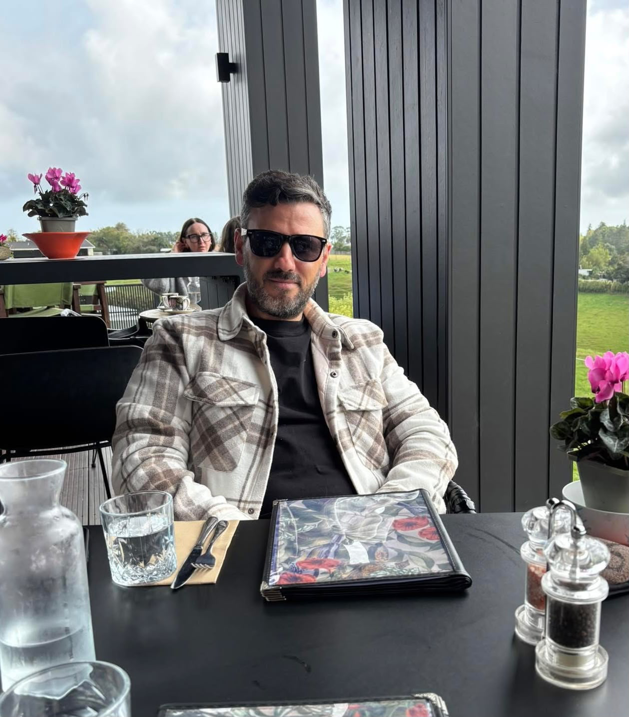 A man with dark hair, beard, and sunglasses sitting at a restaurant table with a black shirt and a plaid jacket, surrounded by flowers and glassware, with a window overlooking green fields and cloudy sky in the background.