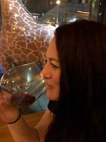 A woman with long dark hair drinking red wine from a glass in a restaurant or bar, with a giraffe statue or decoration visible in the background.