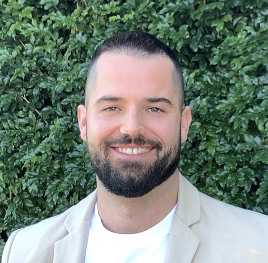 A smiling man with a beard and short dark hair, wearing a cream blazer and white shirt, standing in front of green shrubbery.