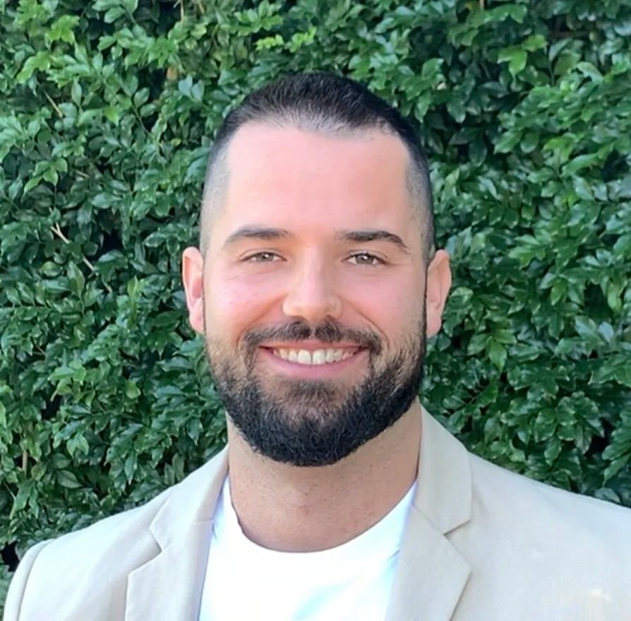 A man with short dark hair and a beard, smiling, wearing a light-colored blazer over a white shirt, standing outdoors in front of green leafy bushes.