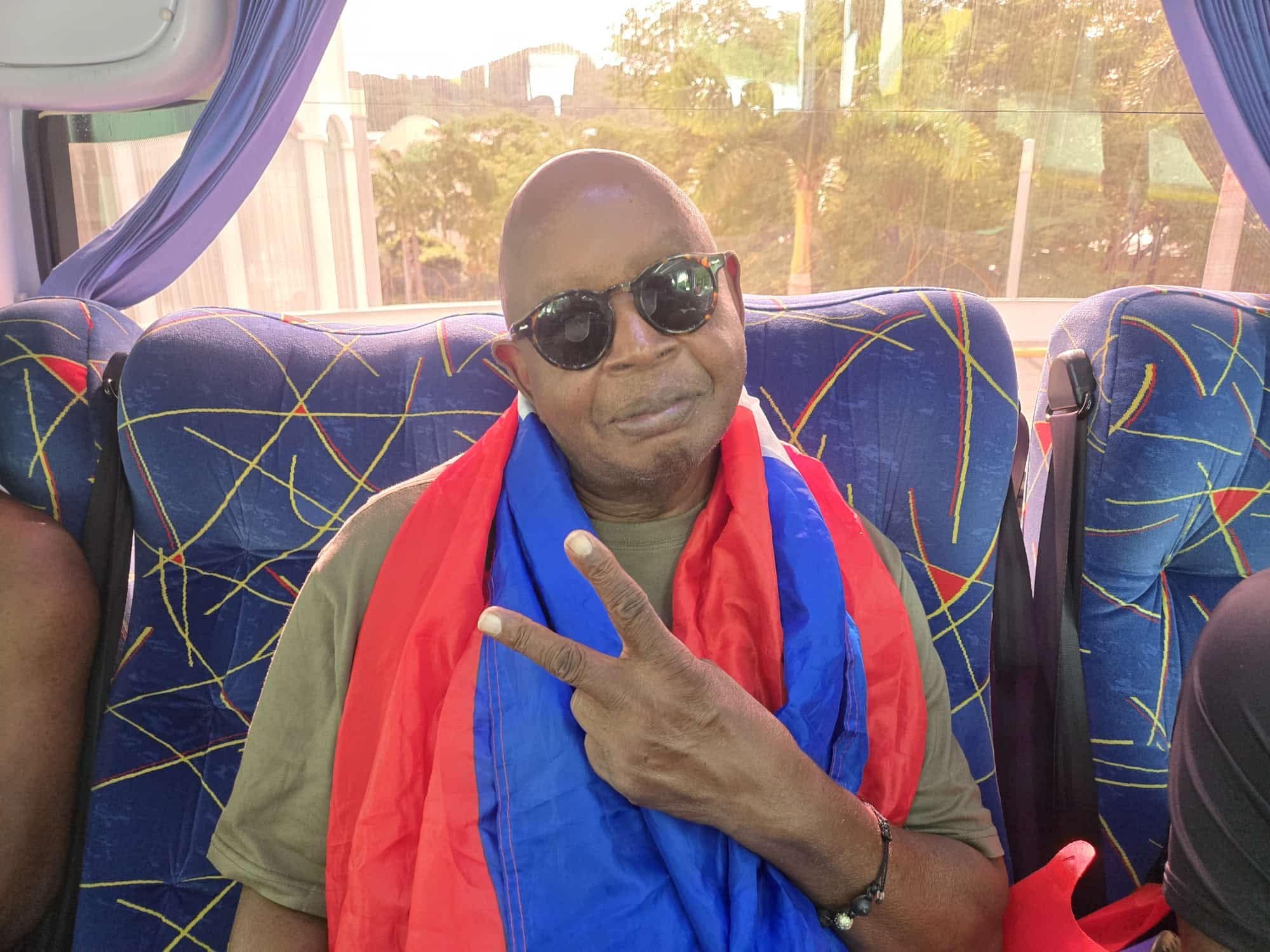 A man sitting on a bus wearing sunglasses, a Nicaragua flag draped over his shoulders, and giving a peace sign gesture.