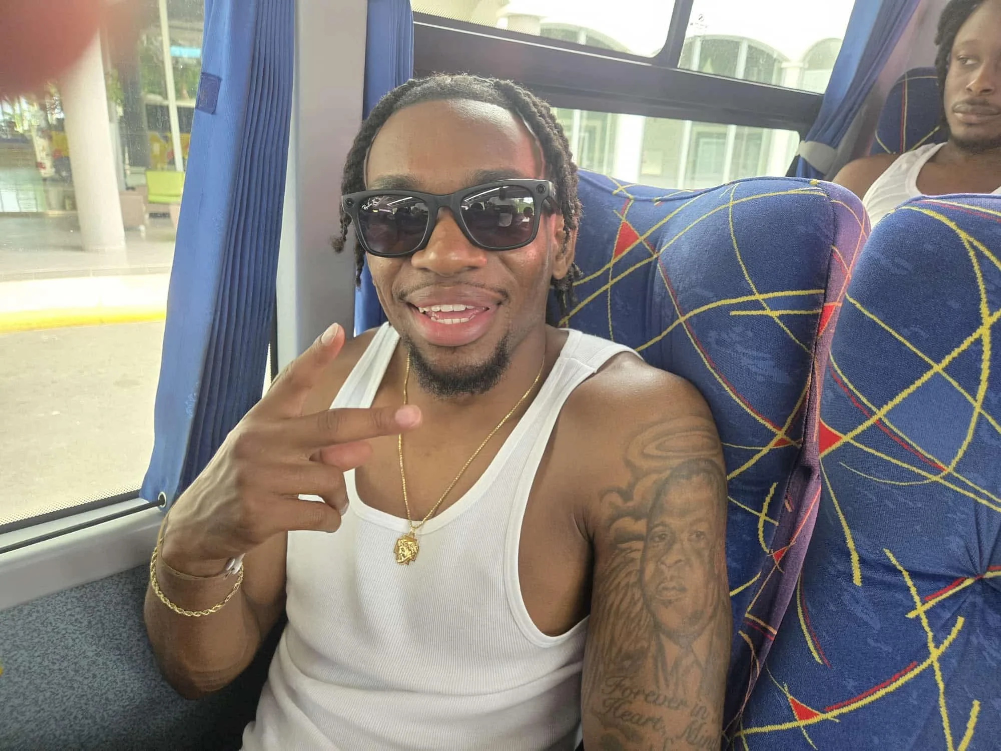 A man with sunglasses taking a selfie inside a bus, pointing with his fingers, wearing a white tank top, gold chain, and tattoos on his arms and chest. Another person is partially visible sitting behind him.