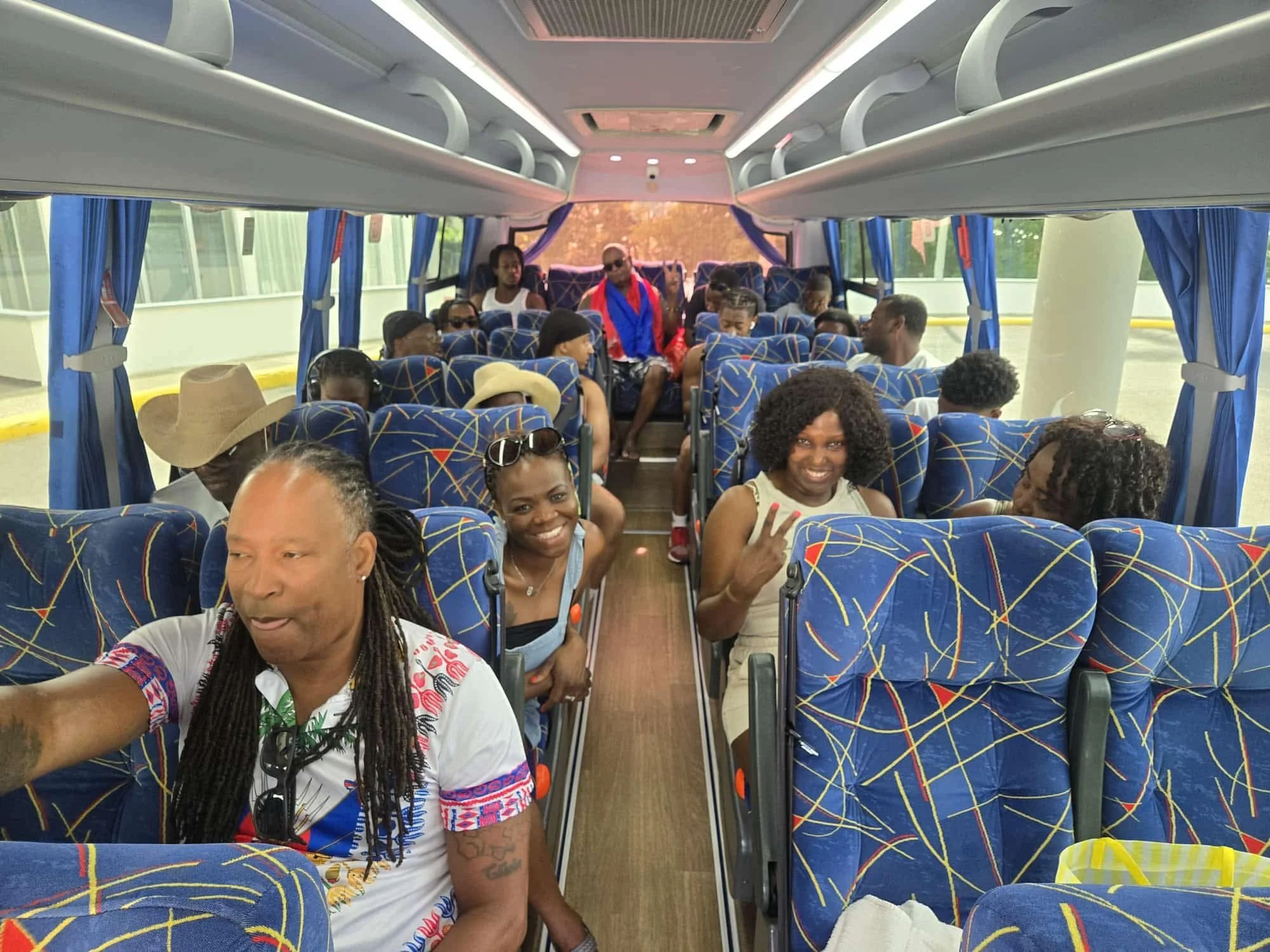 Inside a bus filled with diverse passengers, some smiling and making peace signs, with blue curtains and patterned blue seats.