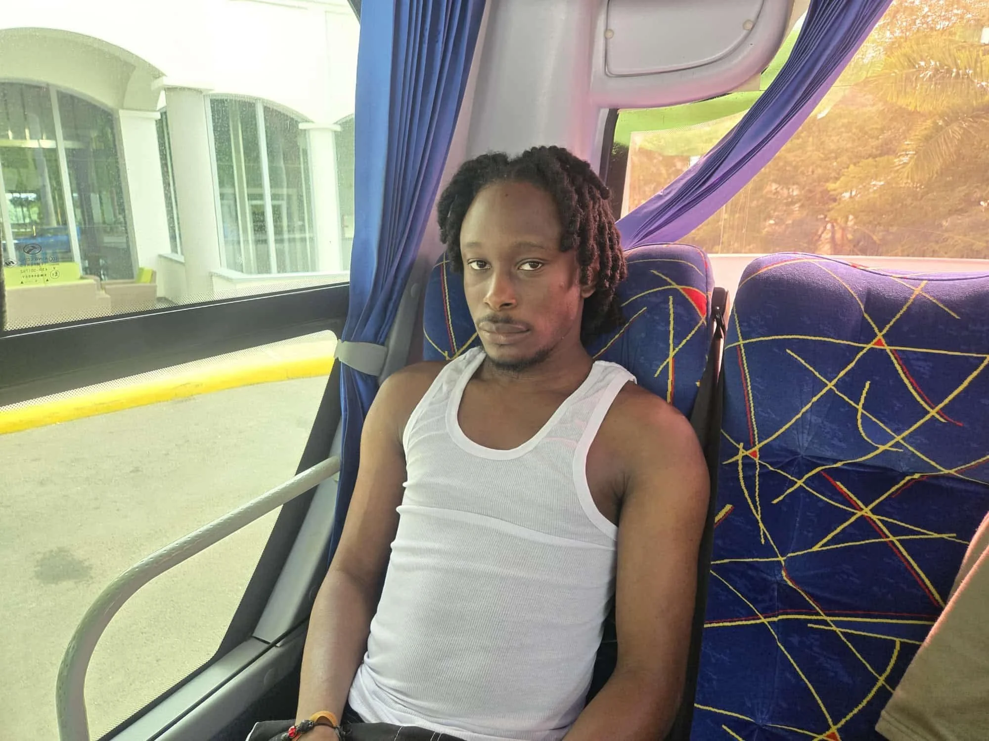 A young man with long dreadlocks, wearing a white sleeveless shirt, sitting on a bus with patterned blue seats. Outside the window, buildings and trees are visible, and the man looks directly at the camera.