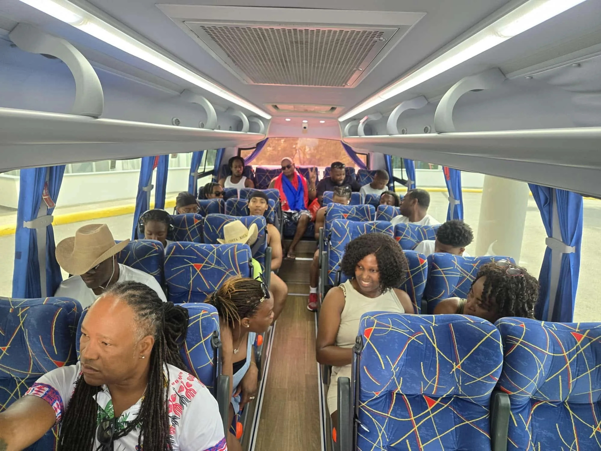 Inside of a bus filled with diverse passengers seated and standing, with some showing peace signs and others engaged in different activities, blue patterned seats, overhead compartments, and windows with blue curtains visible.